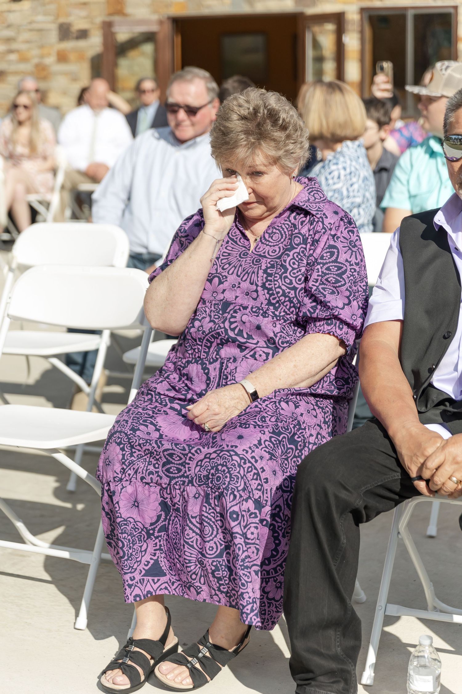 Woman in purple paisley dress sits solemnly at outdoor seating area during daytime event.