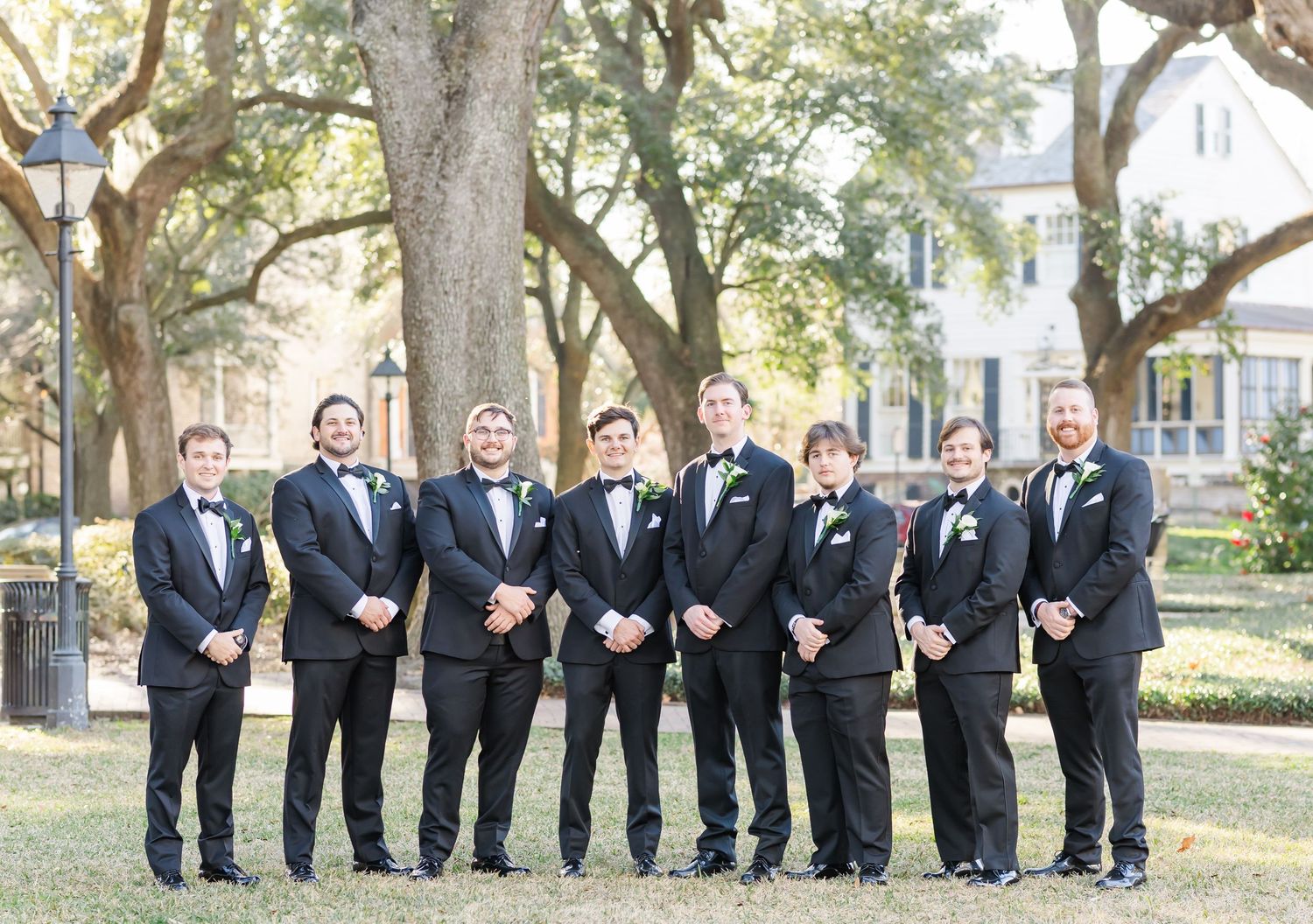 Groomsmen in black tuxedos sitting casually on grass during outdoor wedding photo session.