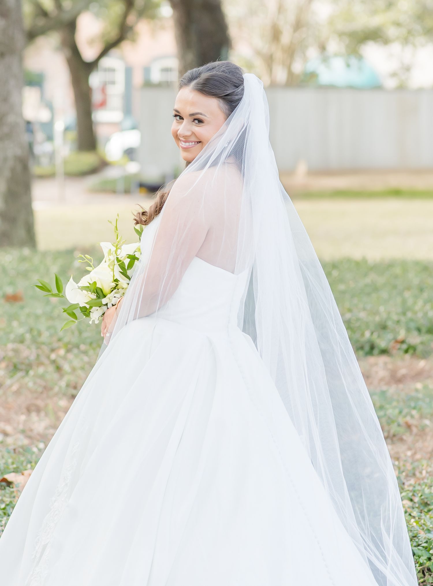 Bride in flowing white wedding dress and veil poses in outdoor garden setting.