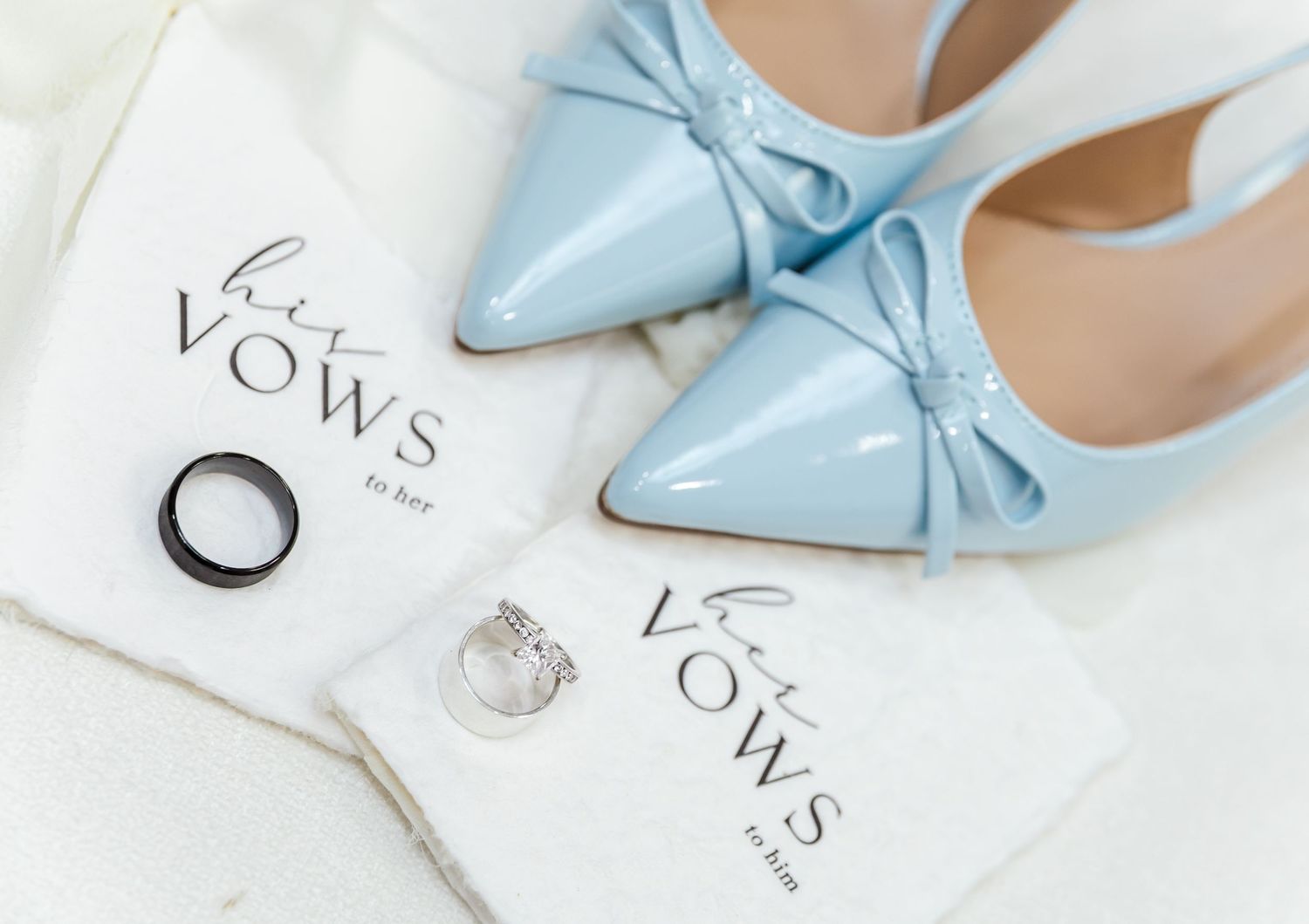 Light blue satin bow pumps with pointed toes sit elegantly on white marble next to wedding rings and invitations.