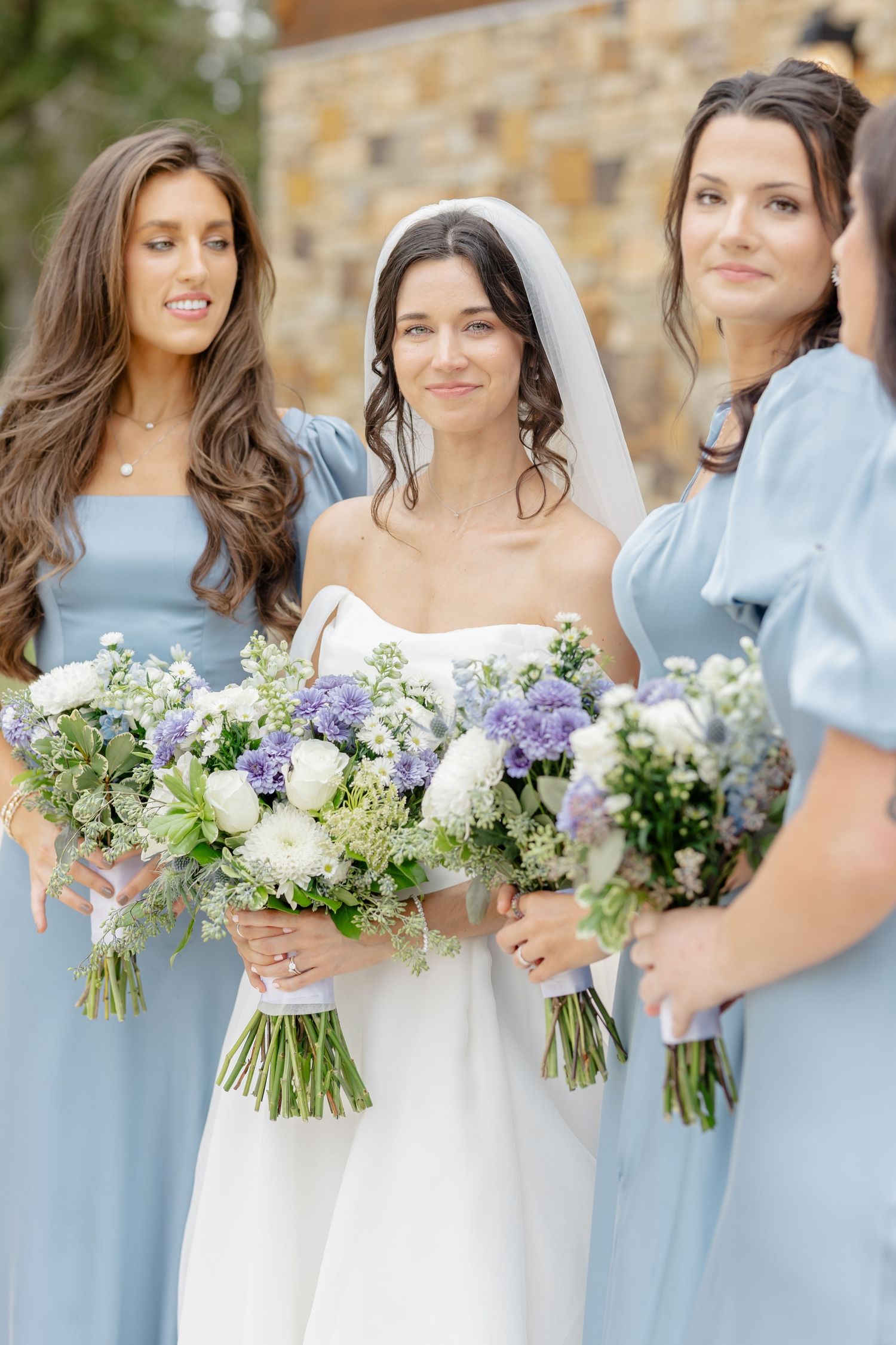 Bridal party holding purple and white flower bouquets pose together in light blue dresses against stone wall.