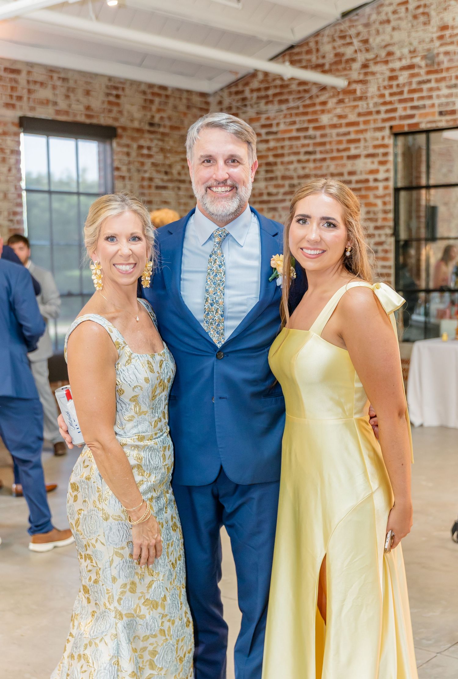 Wedding party members pose in elegant navy suits and pastel yellow and floral dresses against a rustic brick wall backdrop.