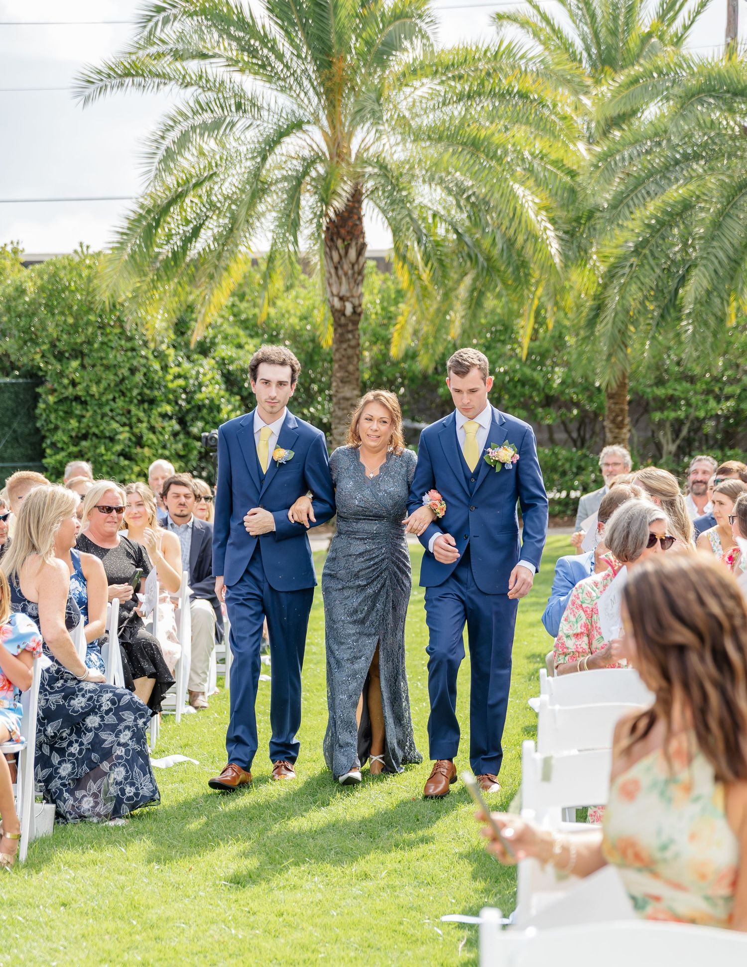 A wedding processional takes place on a lawn with palm trees and guests seated on either side of the aisle.