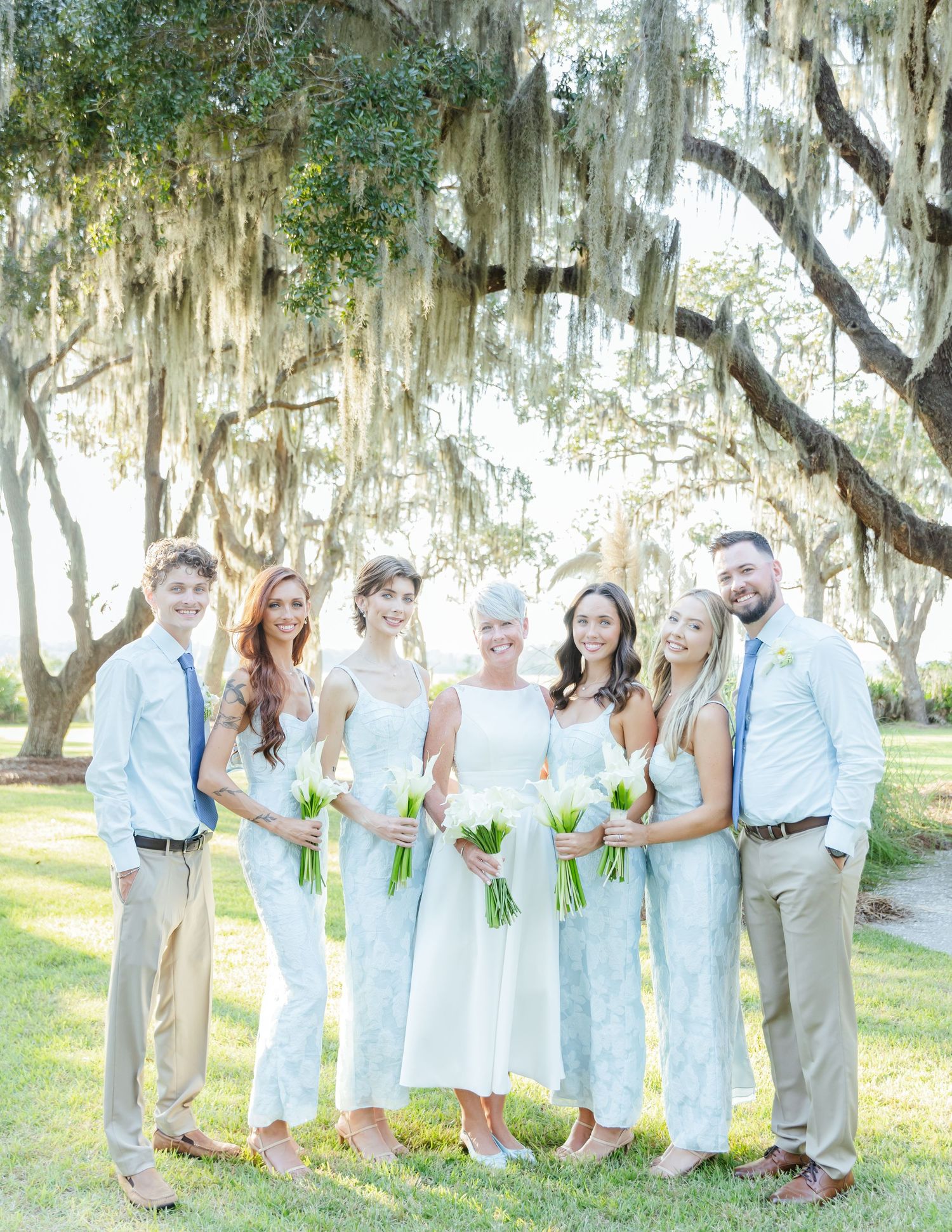 Wedding party in light blue and white attire poses under Spanish moss draped trees in an outdoor setting.
