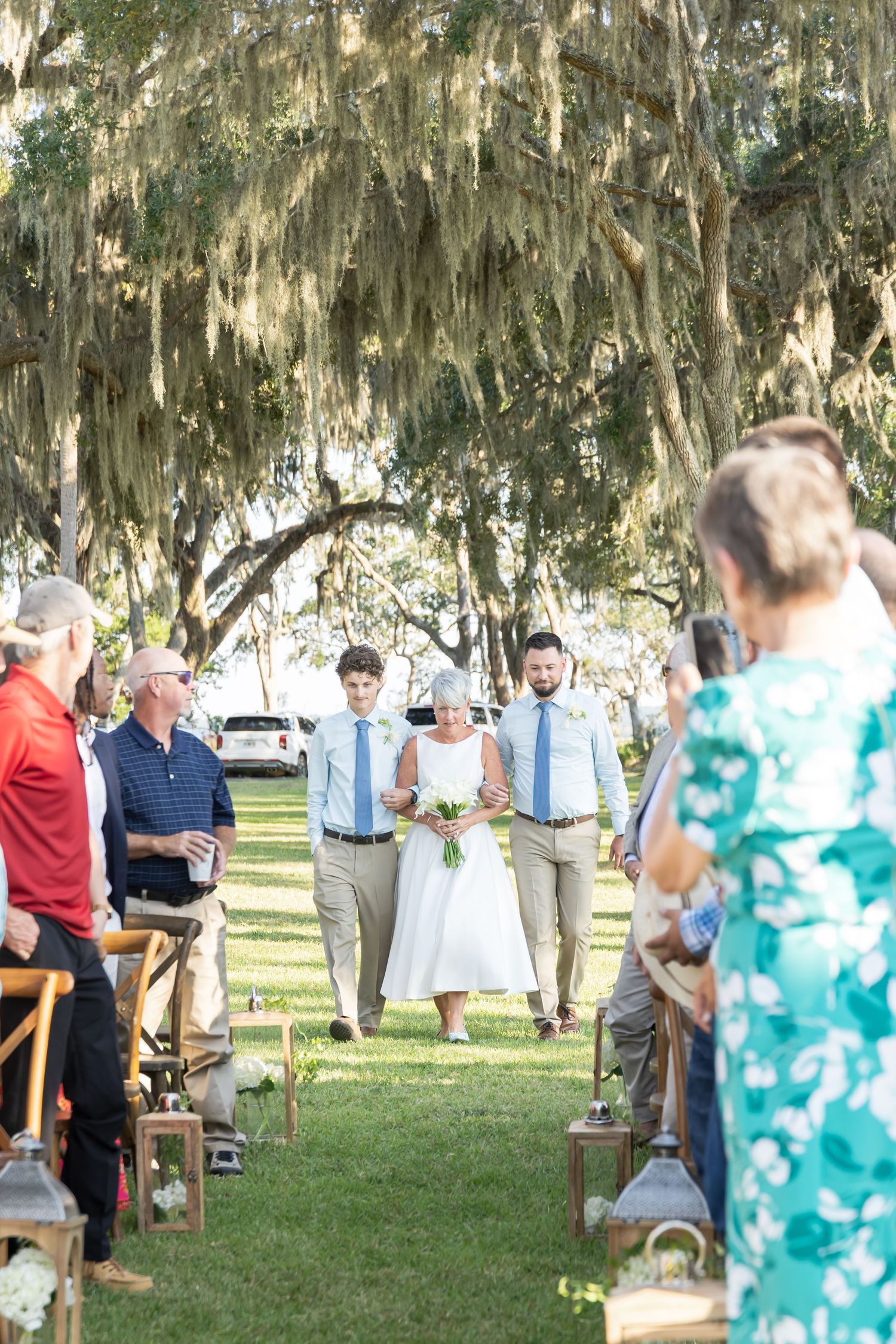 Wedding attendees stand scattered across a sunlit lawn under moss-covered oak trees.
