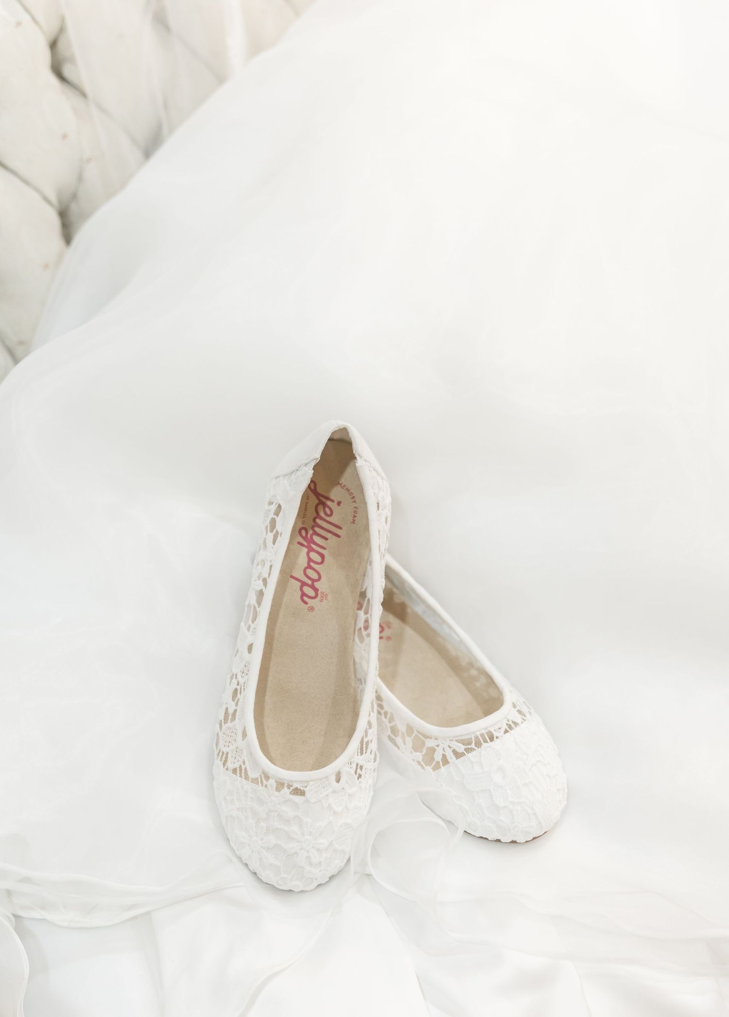 Elegant white lace ballet flats with delicate floral embroidery displayed on soft white fabric background.