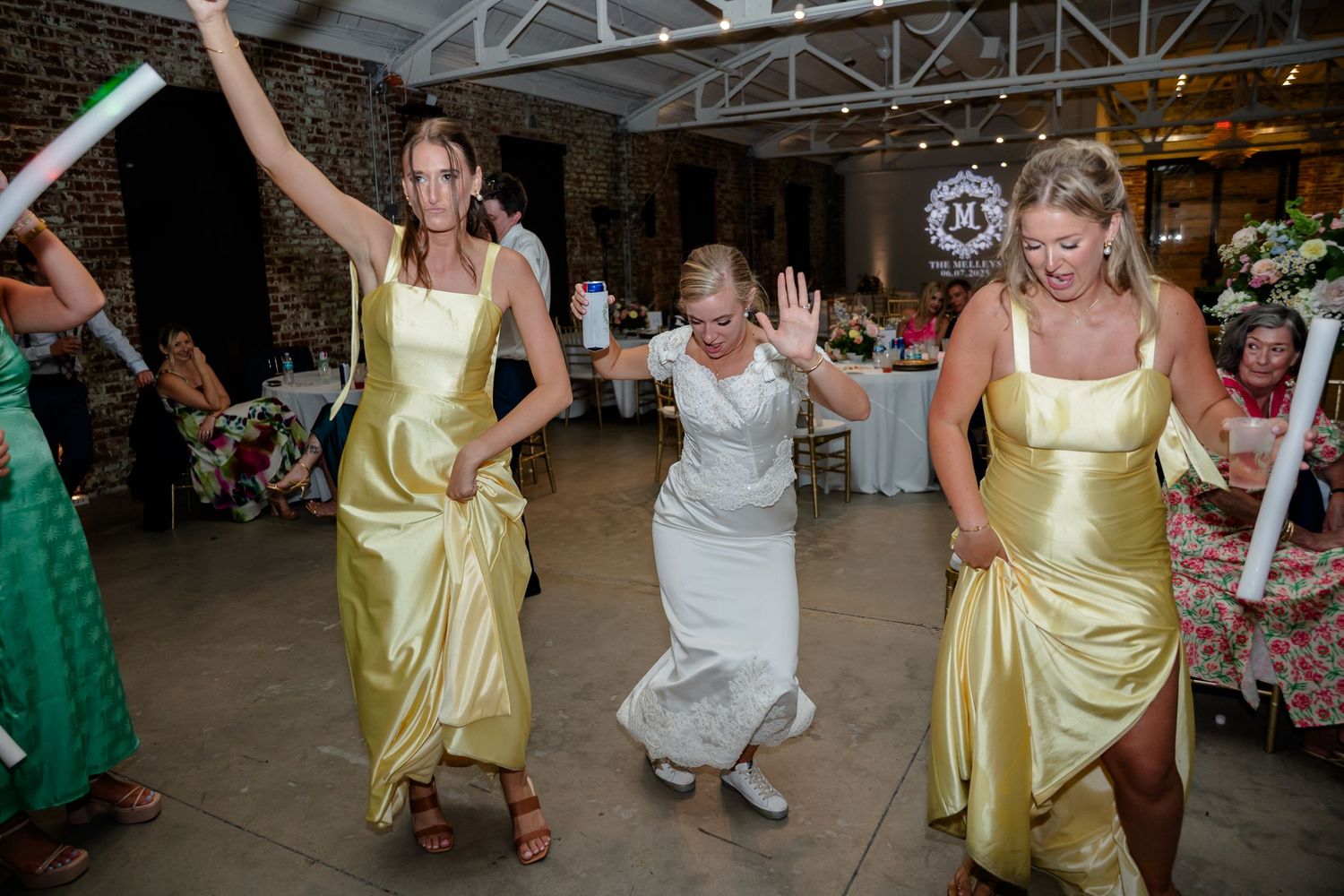 People in yellow and white dresses dance energetically at what appears to be a wedding reception celebration.