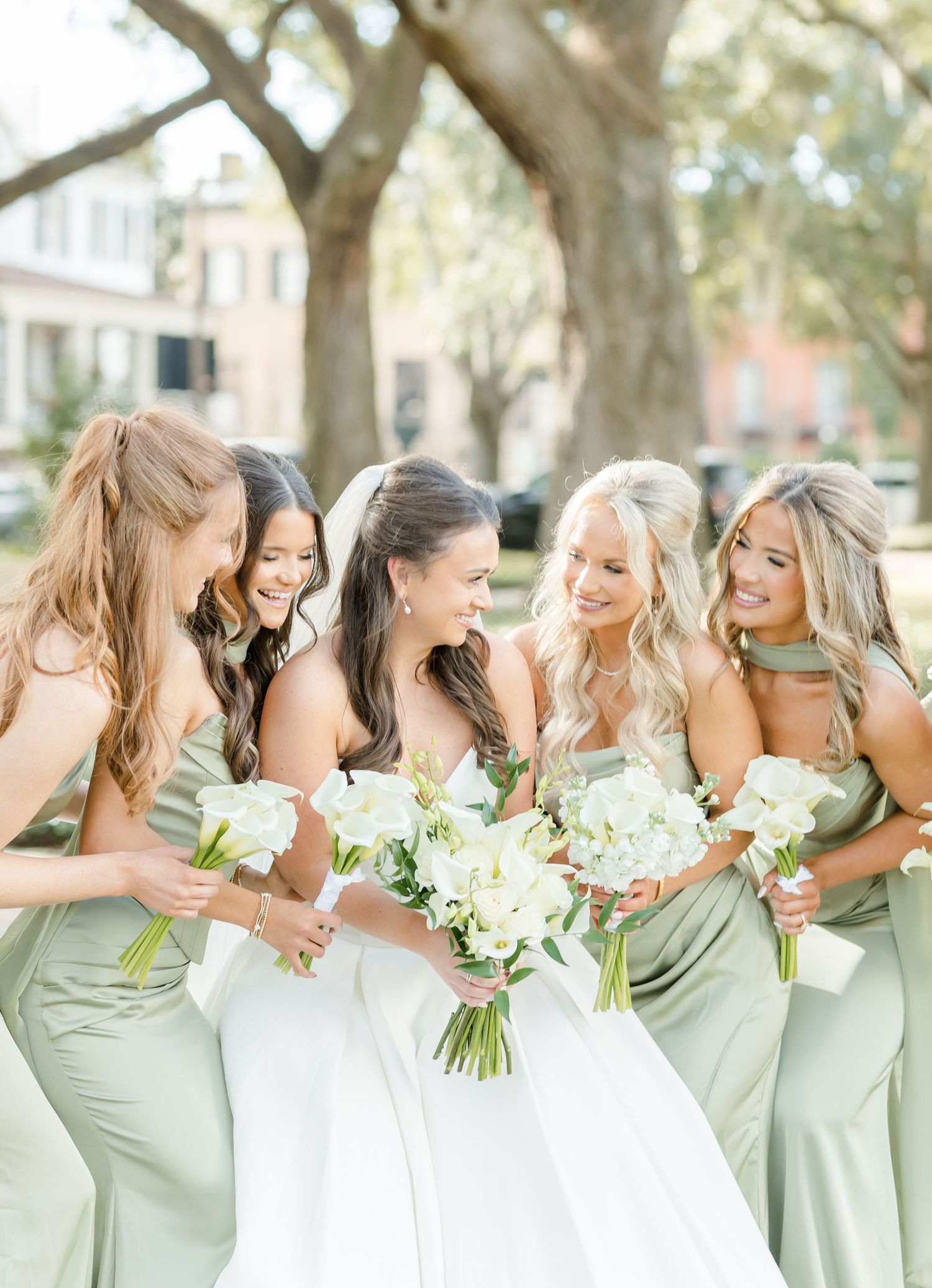 Bridesmaids in sage green dresses share joyful laughter while holding white bouquets during outdoor wedding photoshoot.