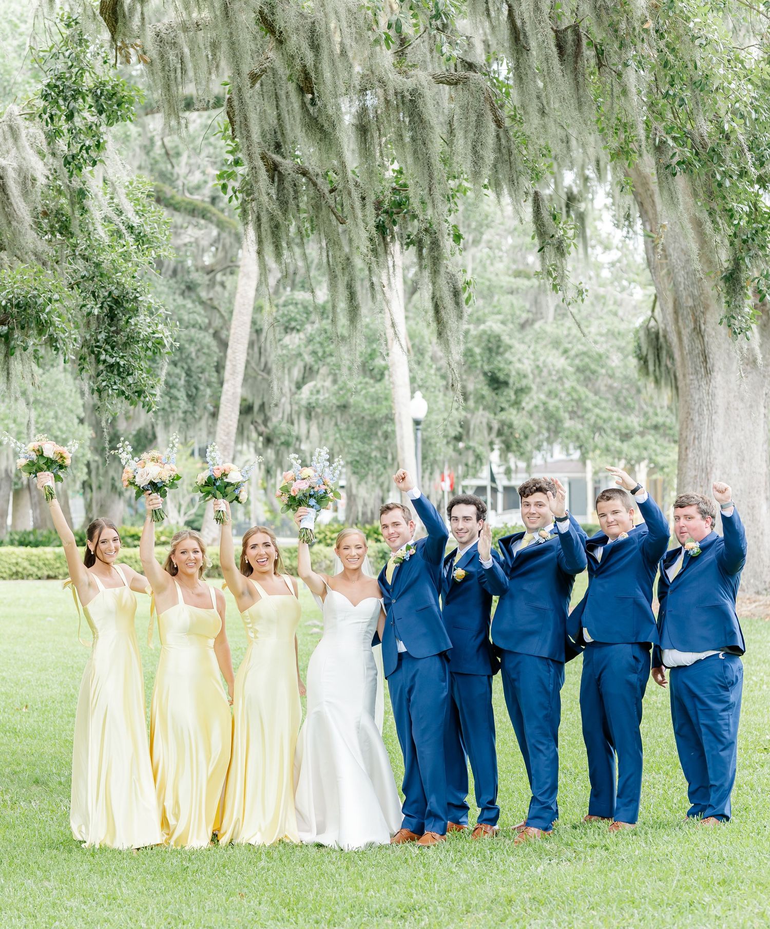 Wedding party wearing navy blue and yellow dresses poses under Spanish moss draped trees in outdoor garden setting.