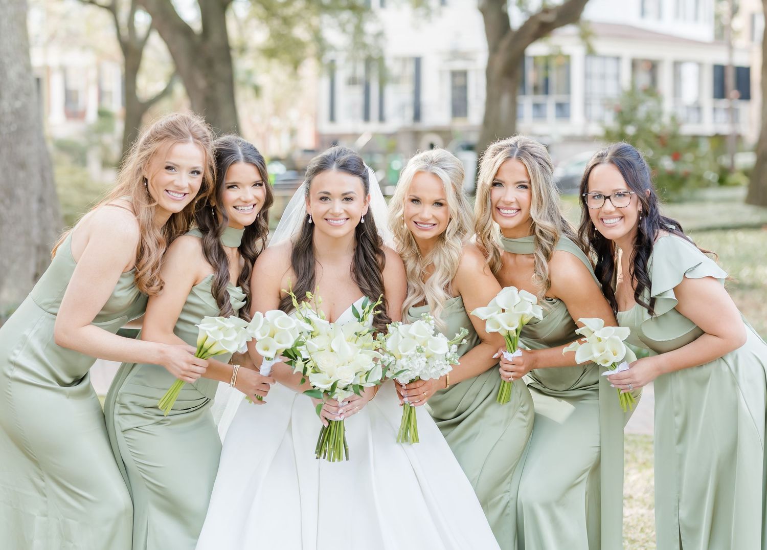 Bride in white gown poses with five bridesmaids in sage green dresses holding white bouquets outdoors.