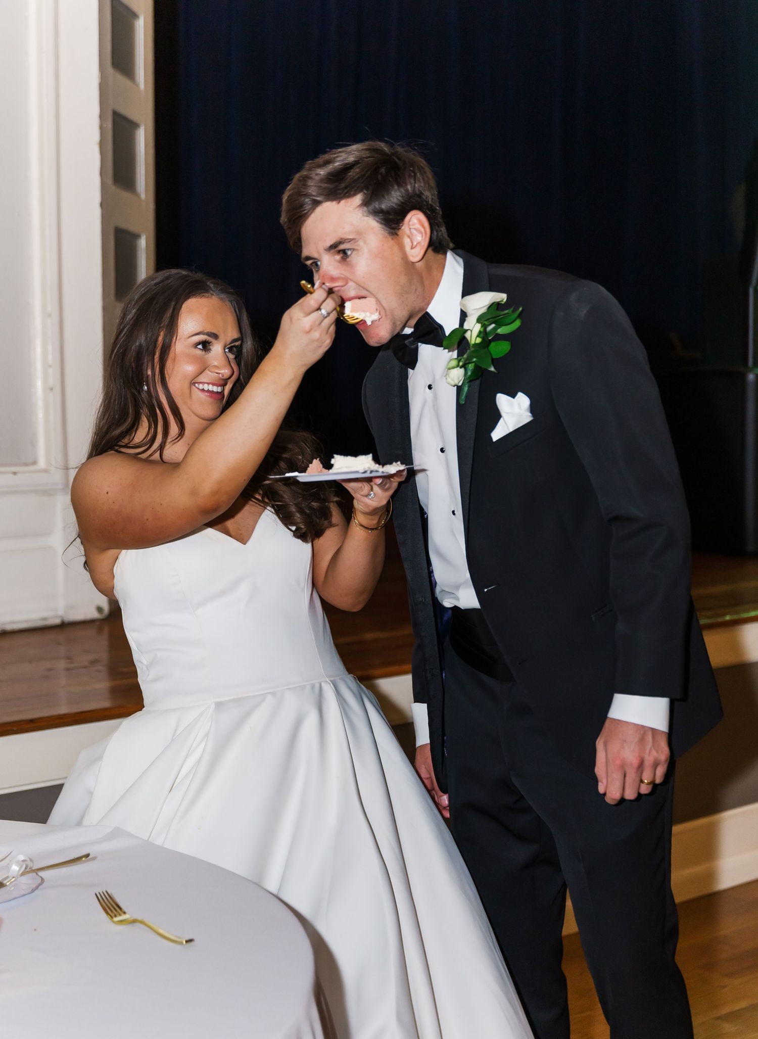 Smiling bride feeding groom a bite of wedding cake at their elegant reception.