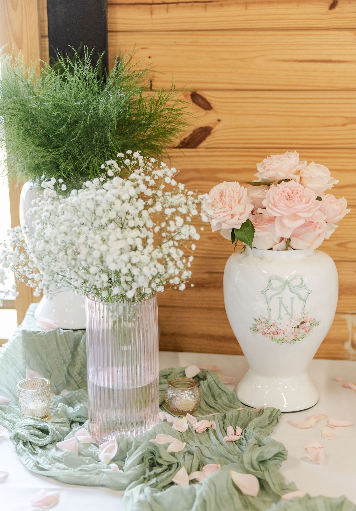 Pink roses in ceramic vase beside baby's breath in glass vase with sage green fabric on white table.