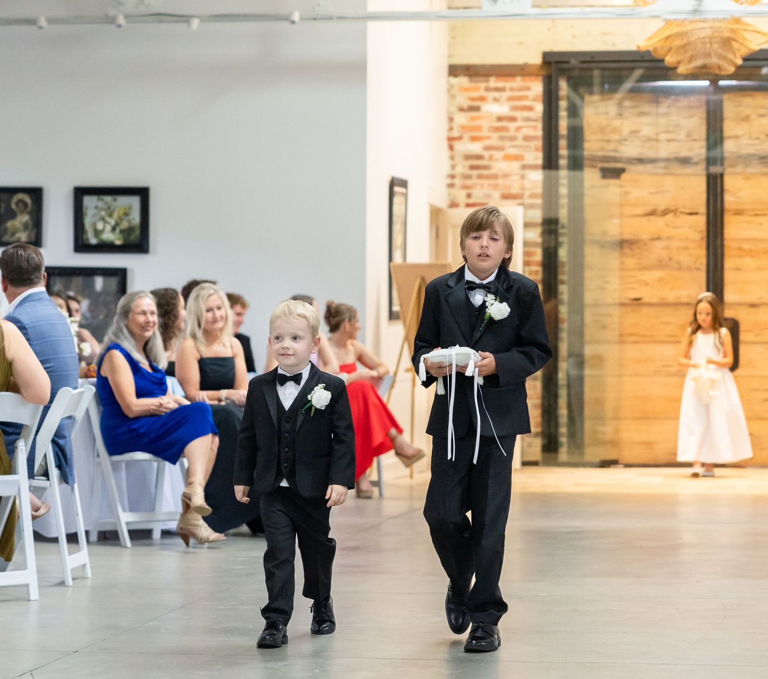 Two young ring bearers in black tuxedos walk down the wedding aisle together during the ceremony.