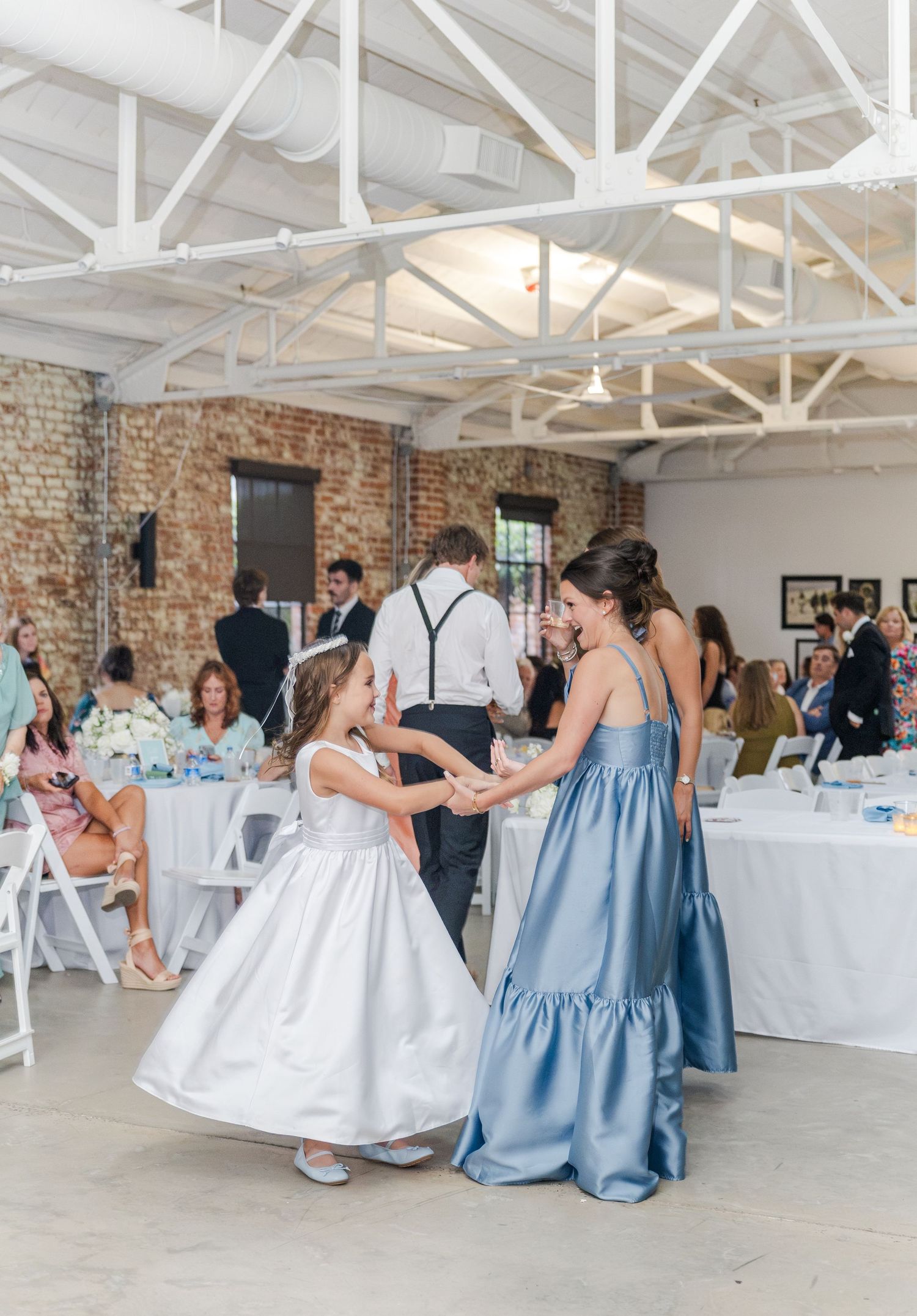 Bridesmaid in blue tiered gown dances with flower girl in white dress at rustic brick reception venue.