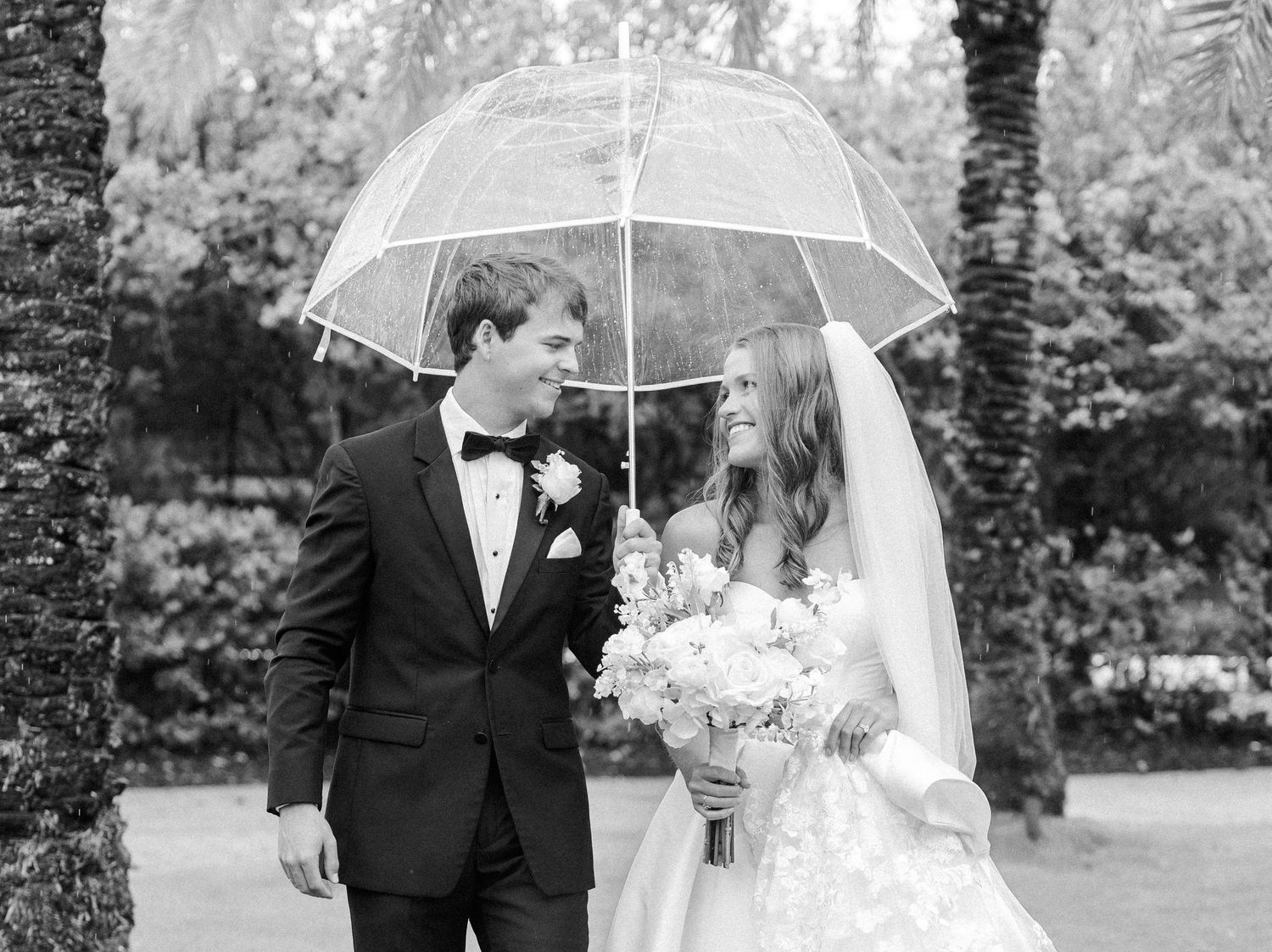 Black and white photo of bride and groom sharing a clear umbrella in the rain on their wedding day.