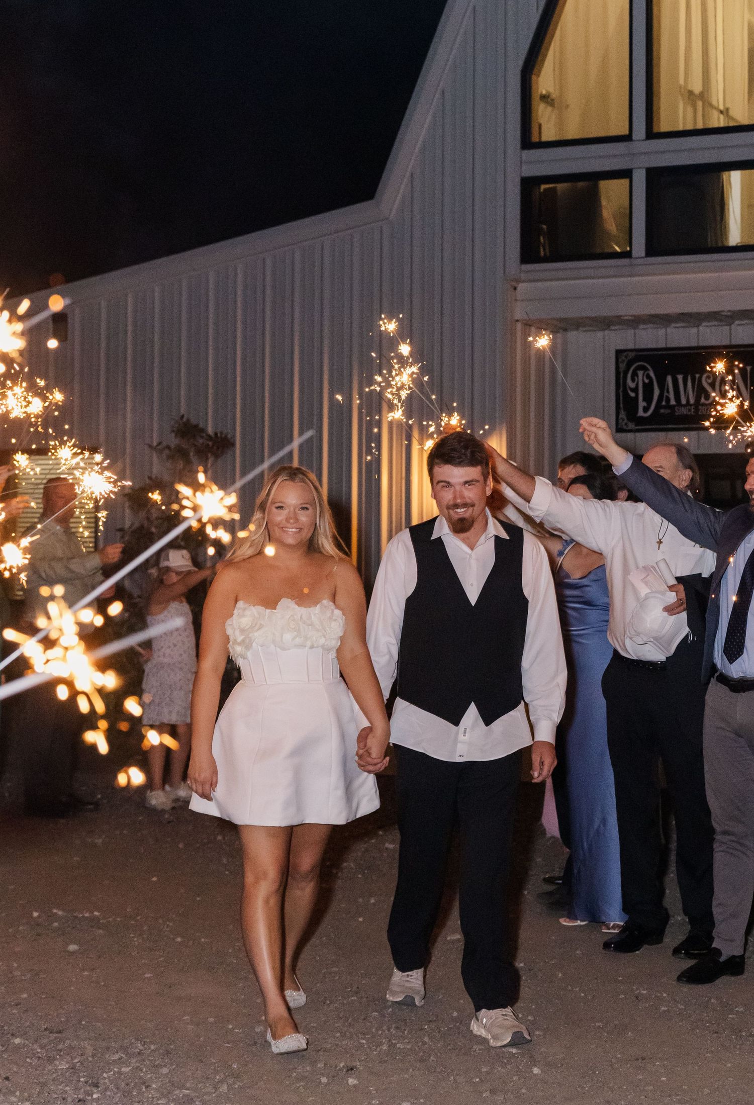 Newlyweds exit wedding reception through sparkler tunnel at night outside white barn venue.