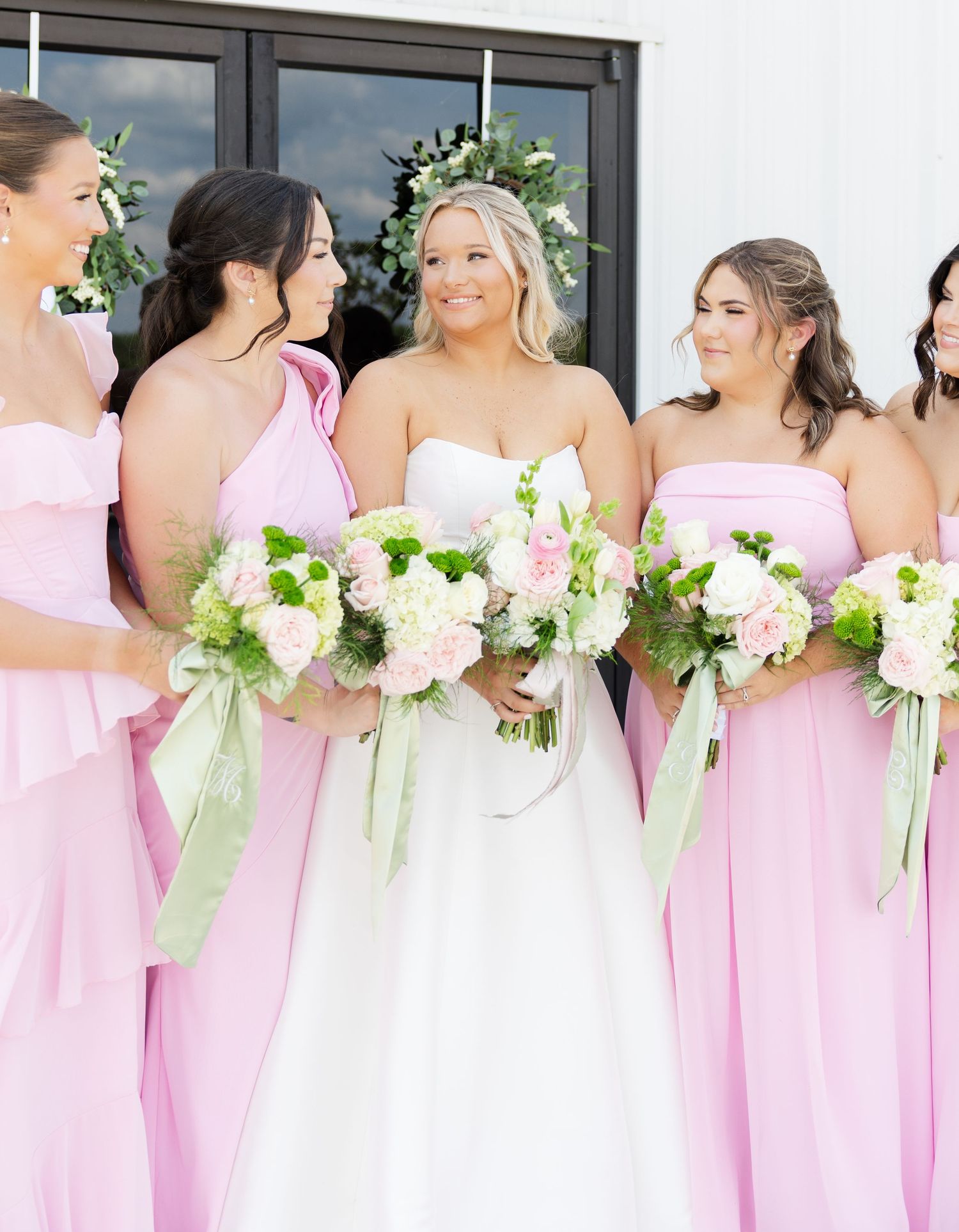 Bride in white strapless gown holds bouquet surrounded by bridesmaids in pink dresses with green-ribboned floral bouquets.