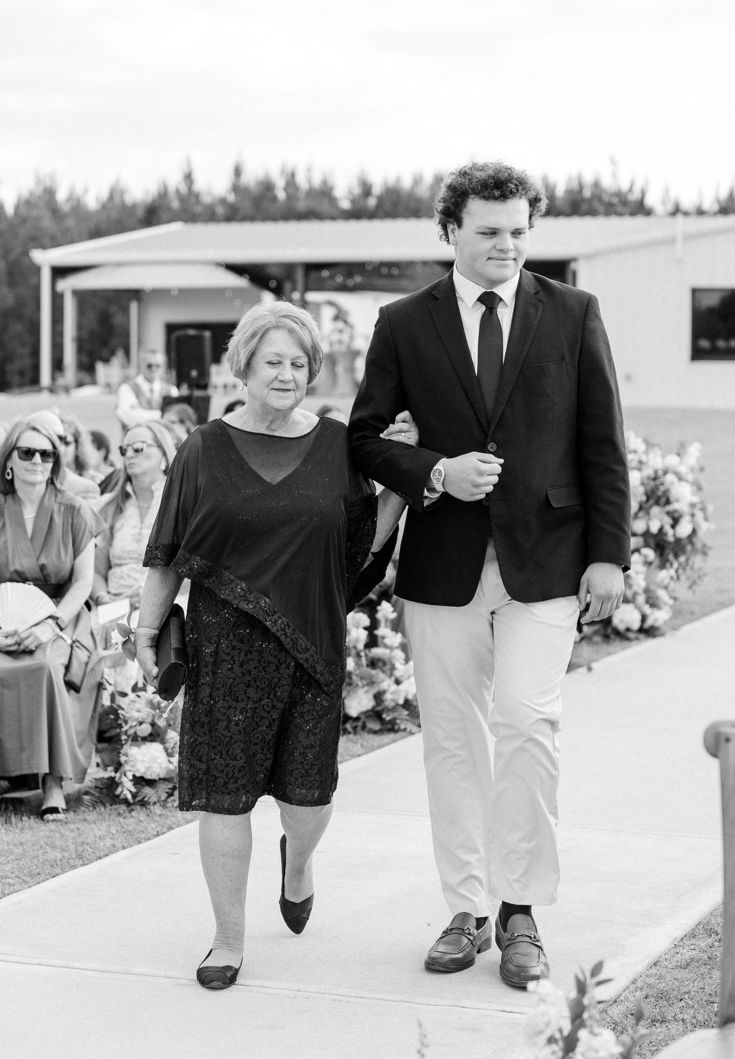 Elegant black and white photo of a young man escorting an older woman down the aisle at an outdoor wedding ceremony.