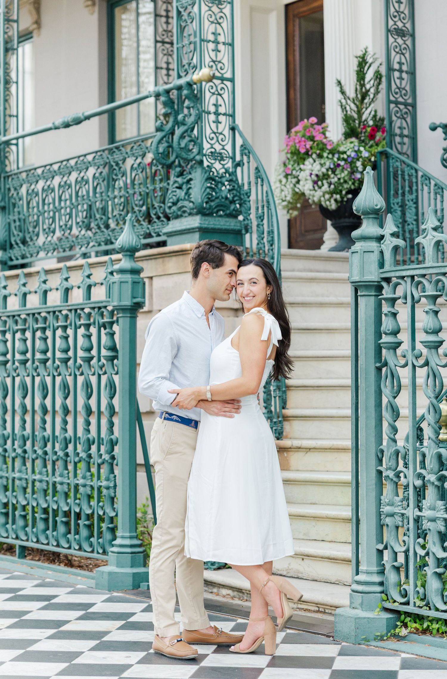 Couple embracing on checkered steps before teal iron railing of historic building with floral decor.