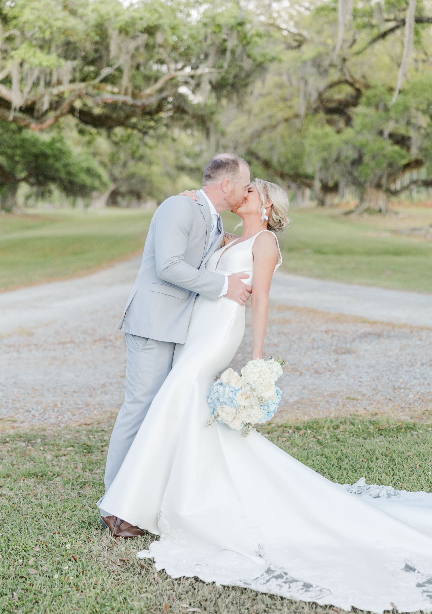 Bride and groom sharing a kiss on a tree-lined path, bride in mermaid gown holding blue and white bouquet.