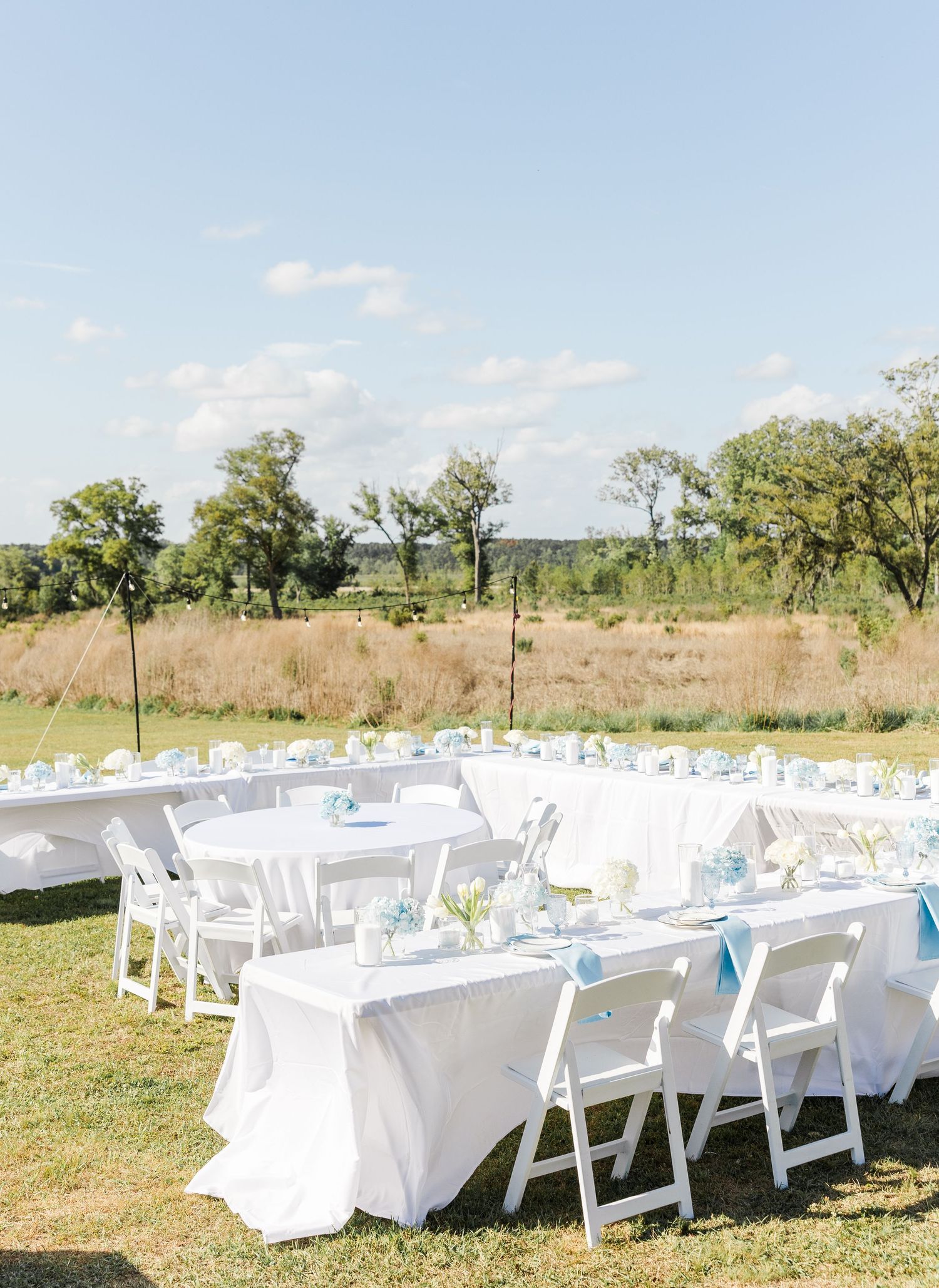 Outdoor wedding reception with white tables, chairs, and blue napkins overlooking open field.