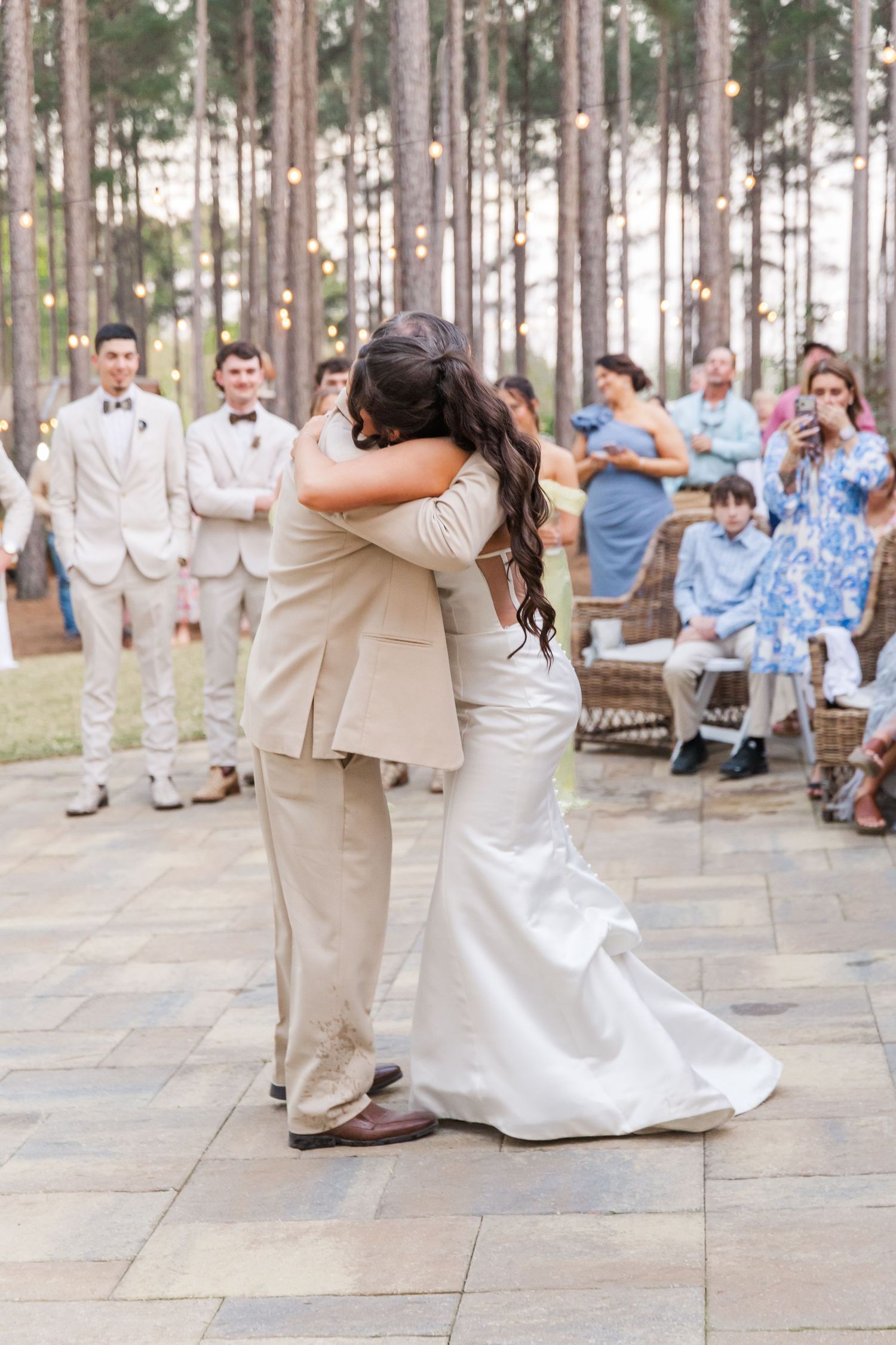 Bride and groom share emotional first dance embrace on stone patio surrounded by guests and string lights.