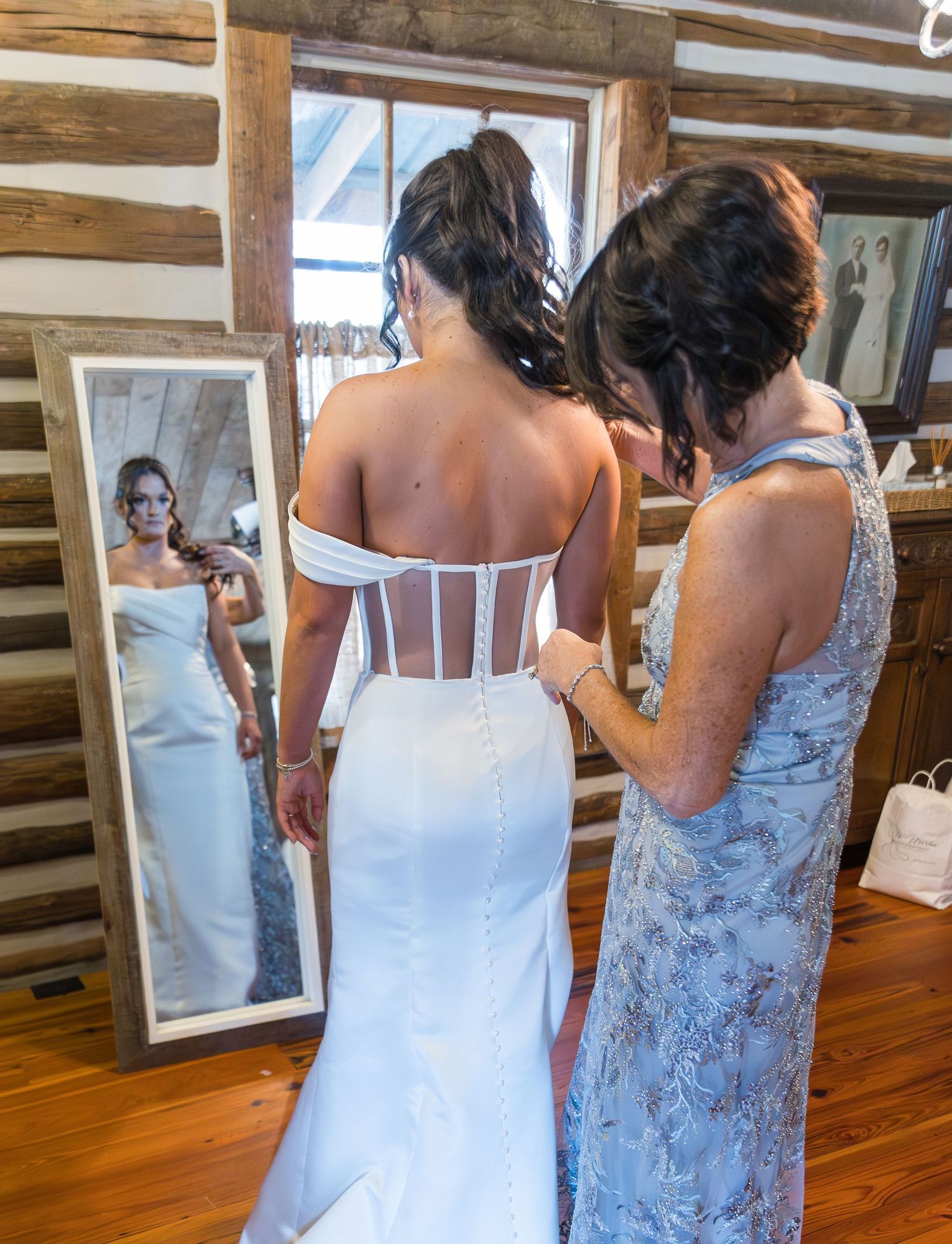 Bride in white corset wedding gown being helped by family member in rustic log cabin with floor mirror reflection.