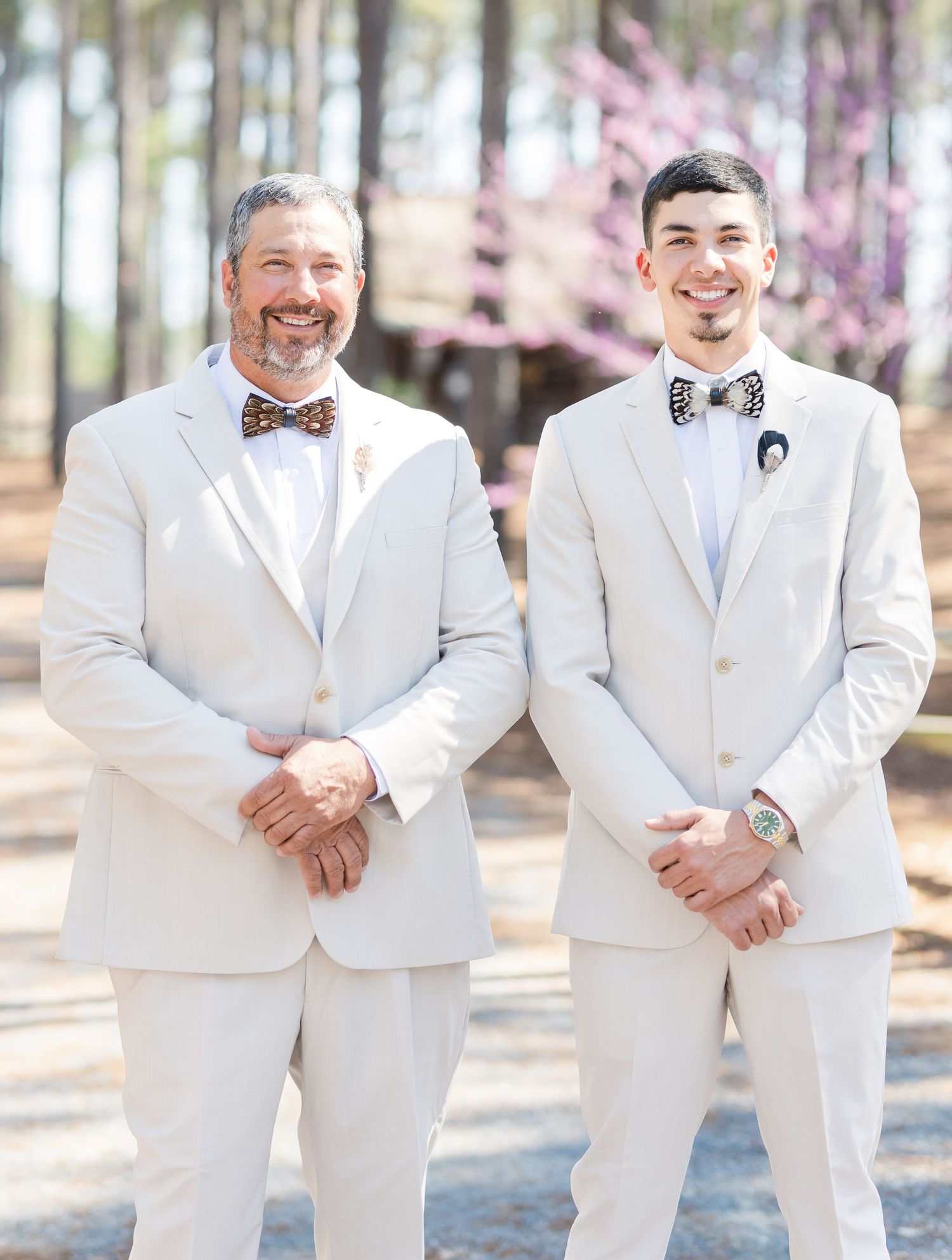 Two groomsmen in cream suits and patterned bow ties smile outdoors at a woodland wedding venue.