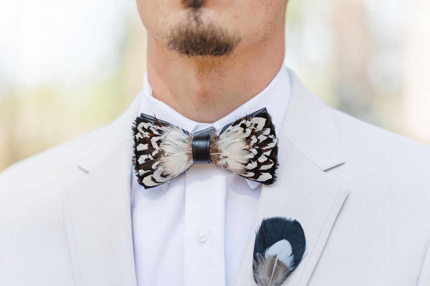 Close-up of groom in white suit jacket wearing a feather bow tie and matching feather pocket square boutonniere.