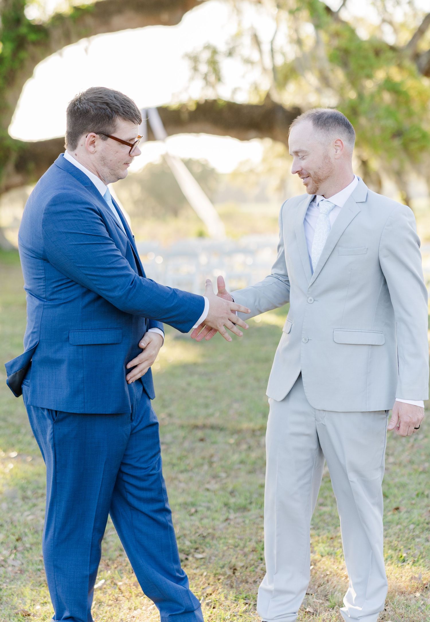 Two groomsmen in blue and gray suits shaking hands outdoors before a wedding ceremony.