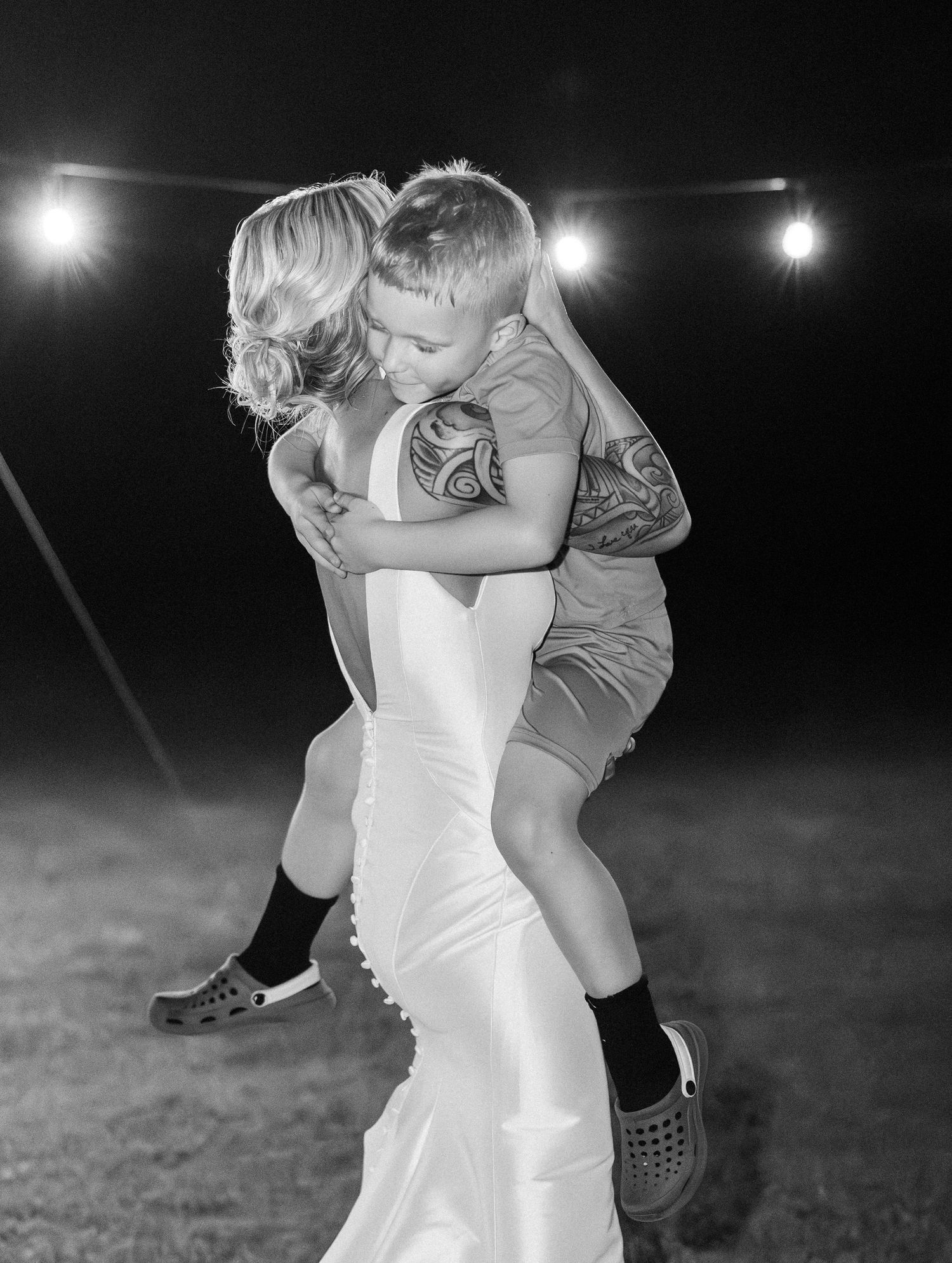 Black and white photo of bride in satin gown hugging a young boy under string lights at night.