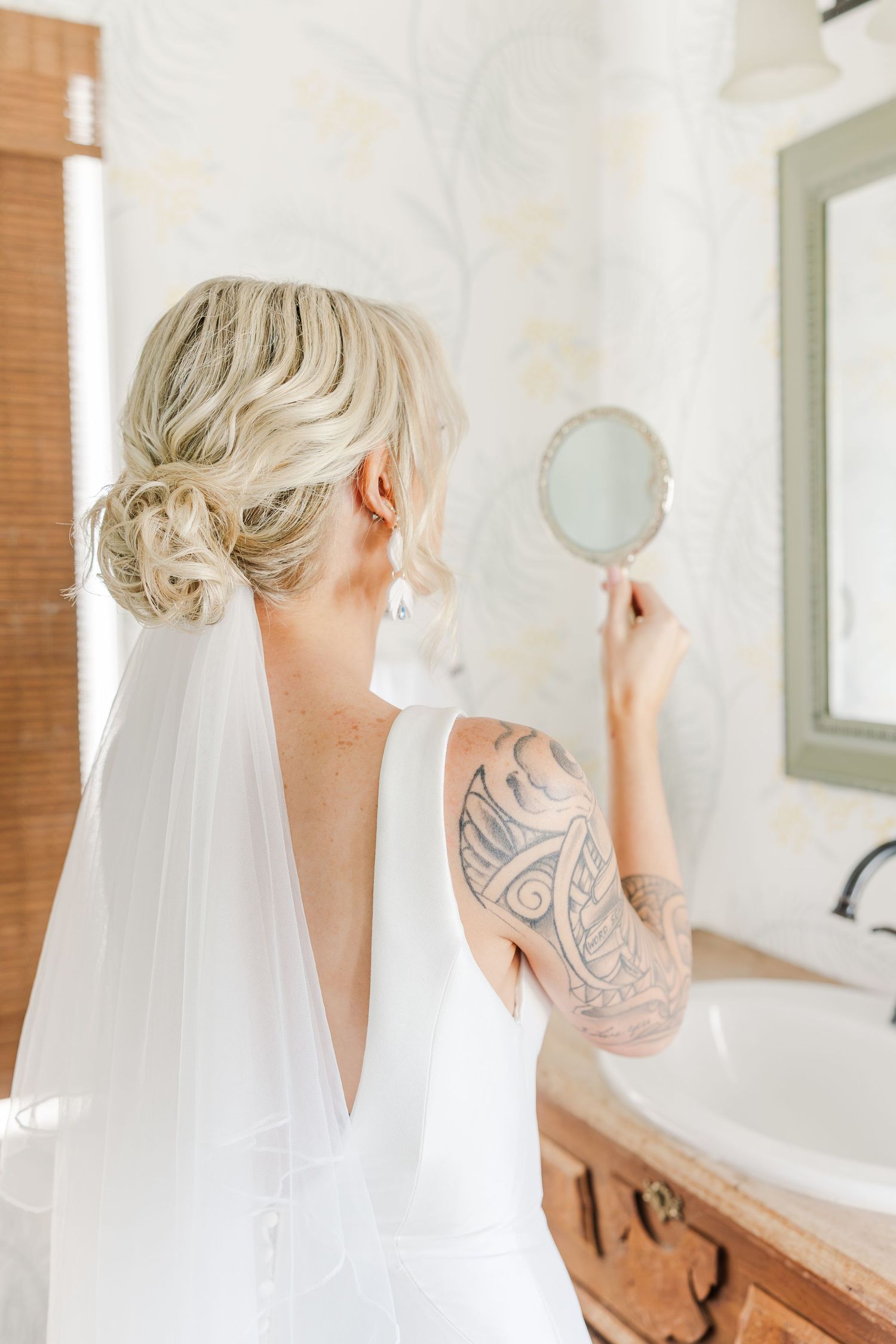 Tattooed blonde bride in backless white gown with veil checks hair in handheld mirror in bathroom.