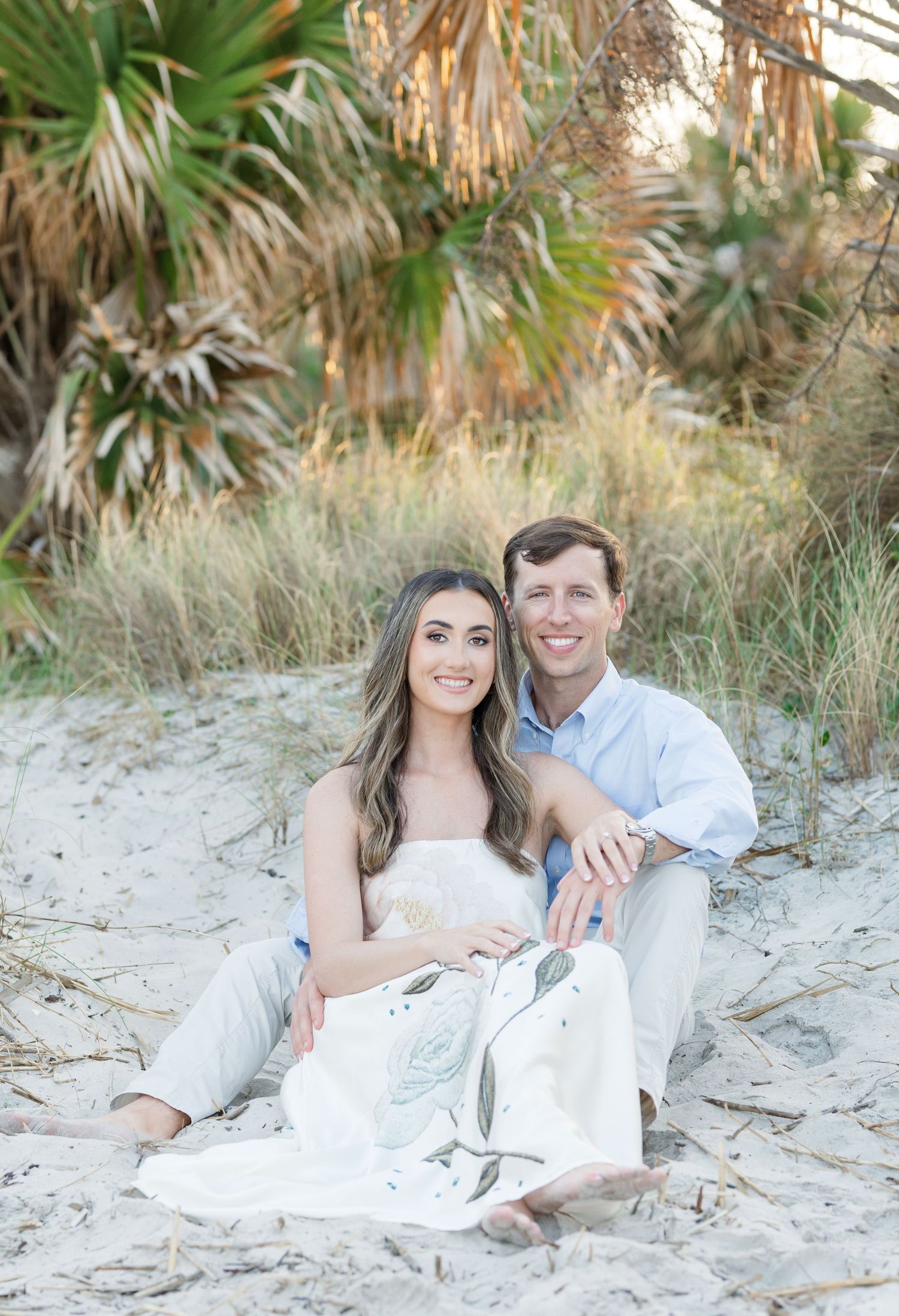 Couple sitting on sandy beach dunes smiling, surrounded by lush palm trees and sea grass.