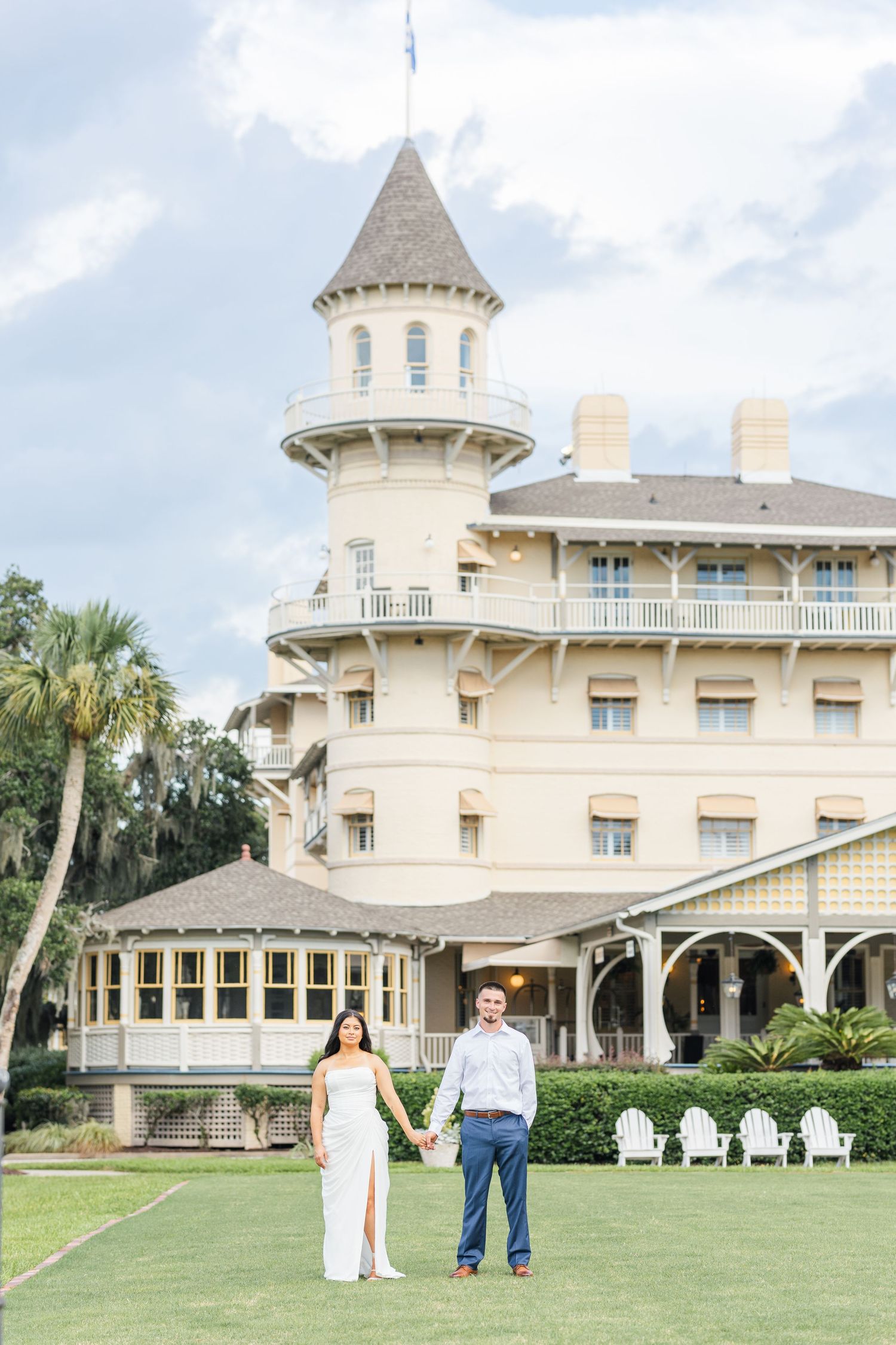 Couple holds hands on lawn before grand Victorian yellow hotel with turret and wrap-around porch.