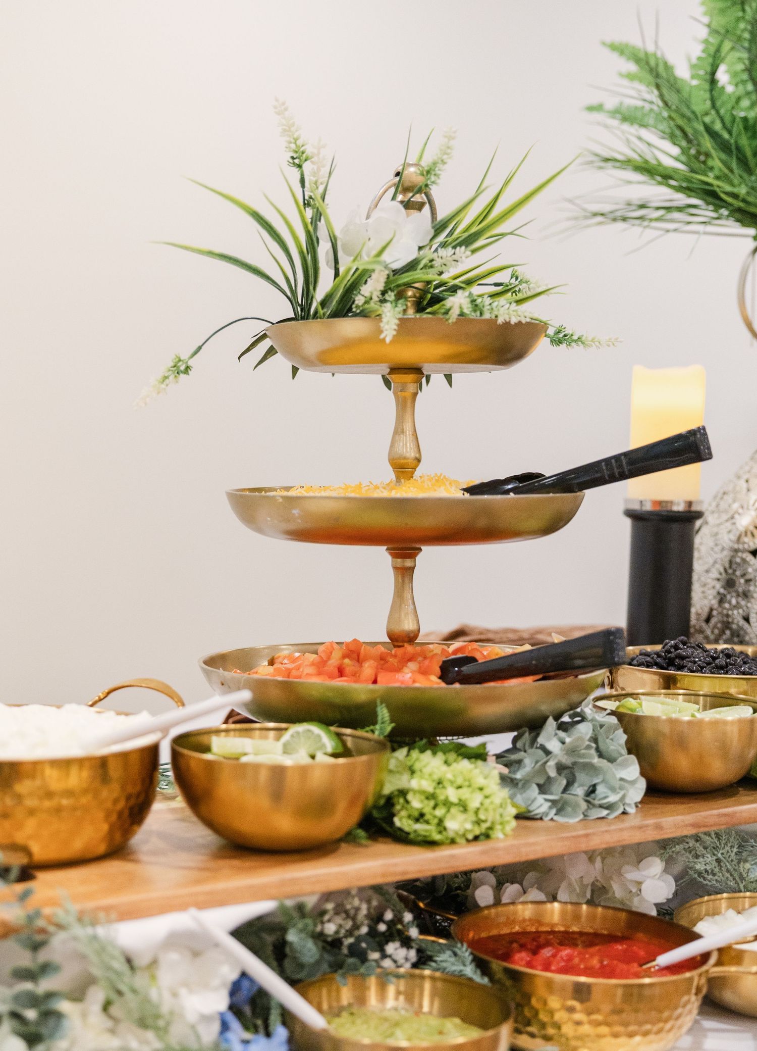 Elegant three-tier gold stand with fresh greenery atop, surrounded by brass bowls filled with colorful food at a buffet.
