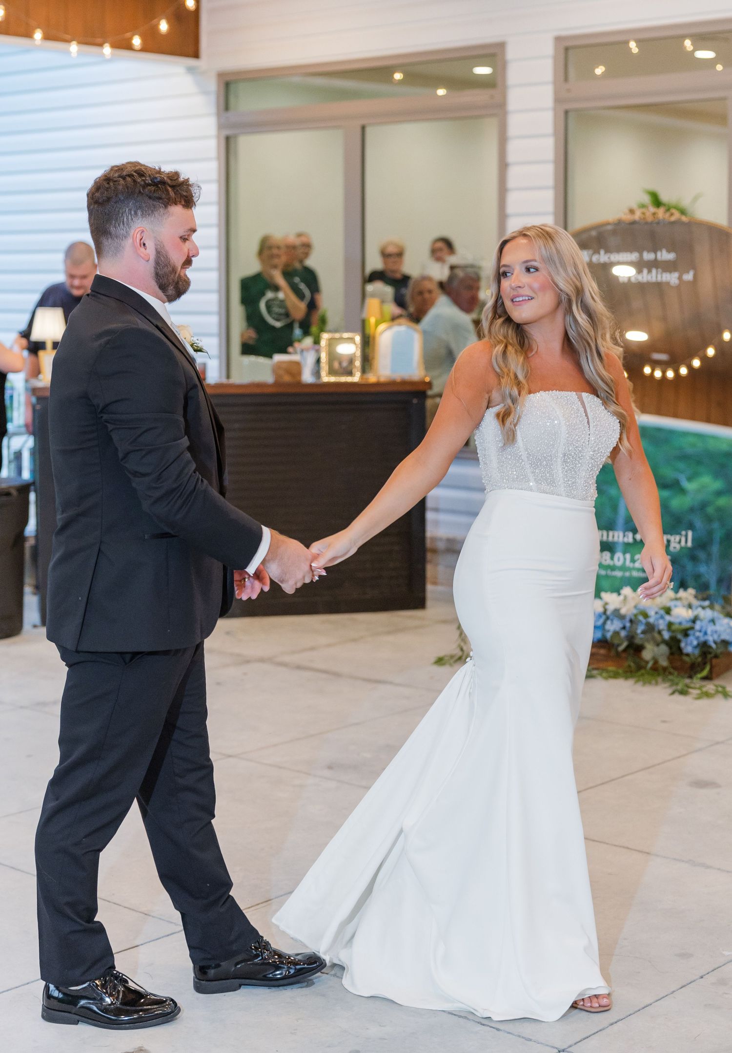 Bride in fitted white gown and groom in black suit hold hands at wedding reception entrance.