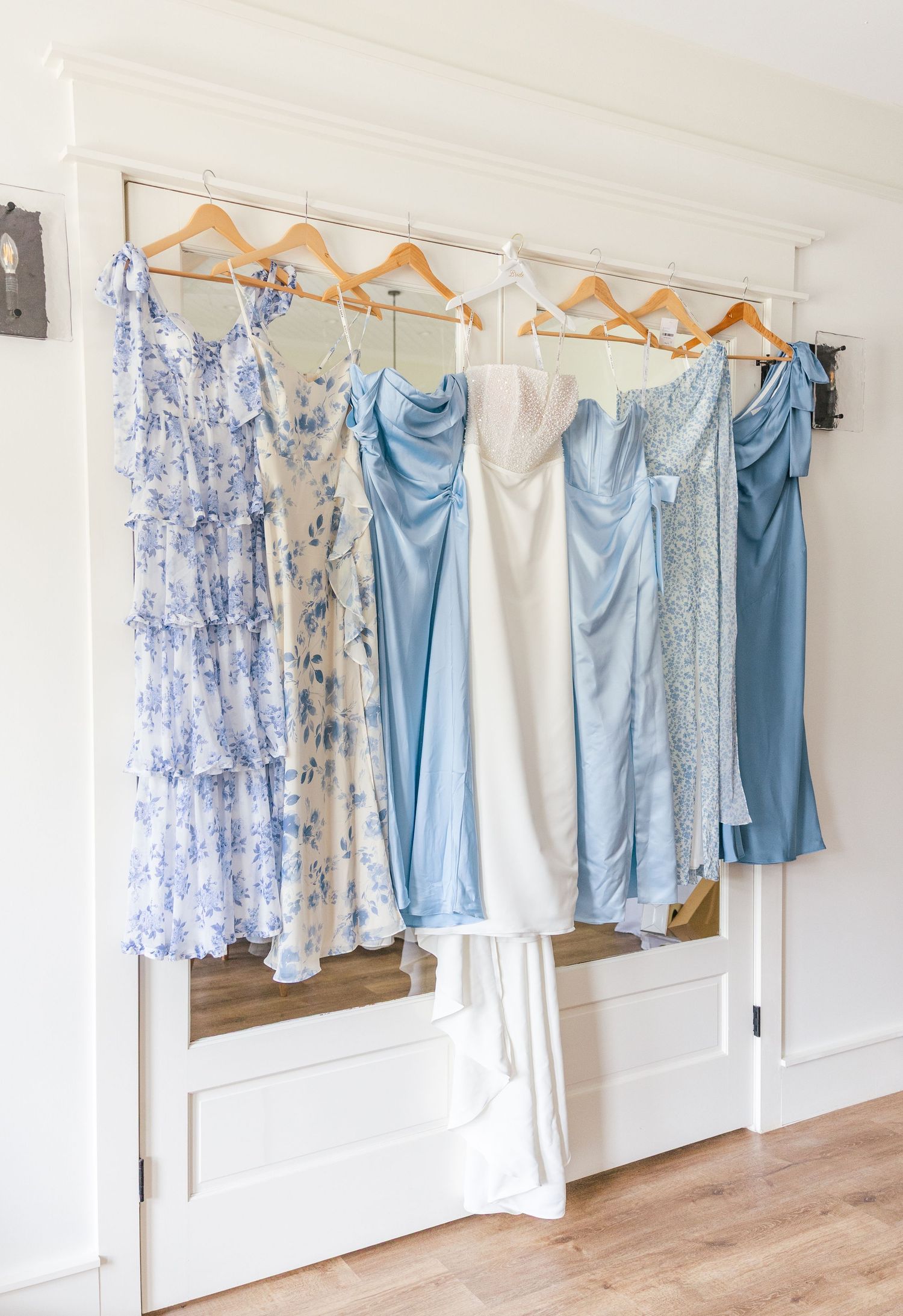Blue and white bridesmaid dresses hanging on wooden hangers against a white wall for a wedding.