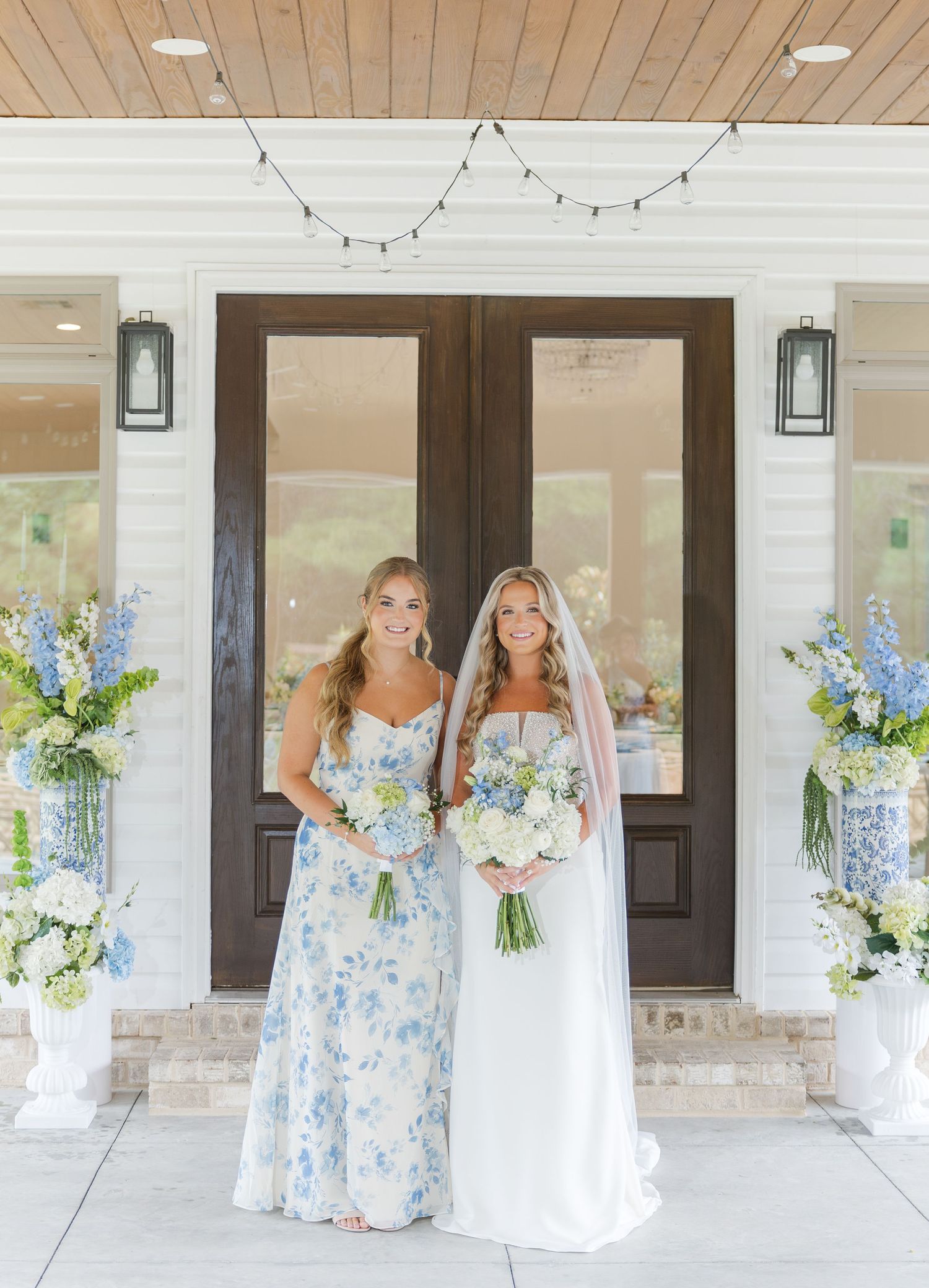 Bride in white gown and bridesmaid in blue floral dress pose at wedding venue entrance.