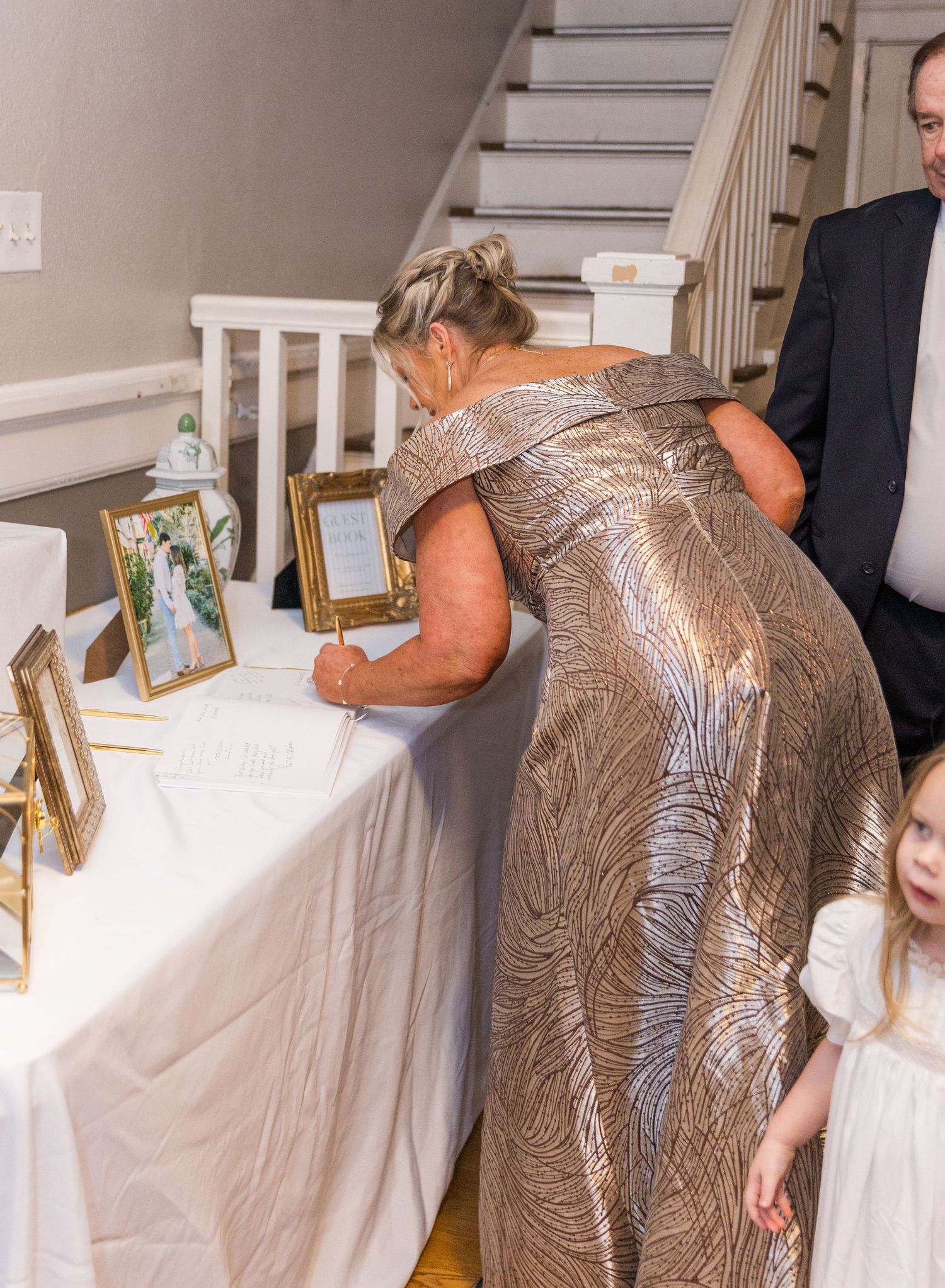 Woman in gold gown signing guest book at elegant wedding reception table with framed photos.