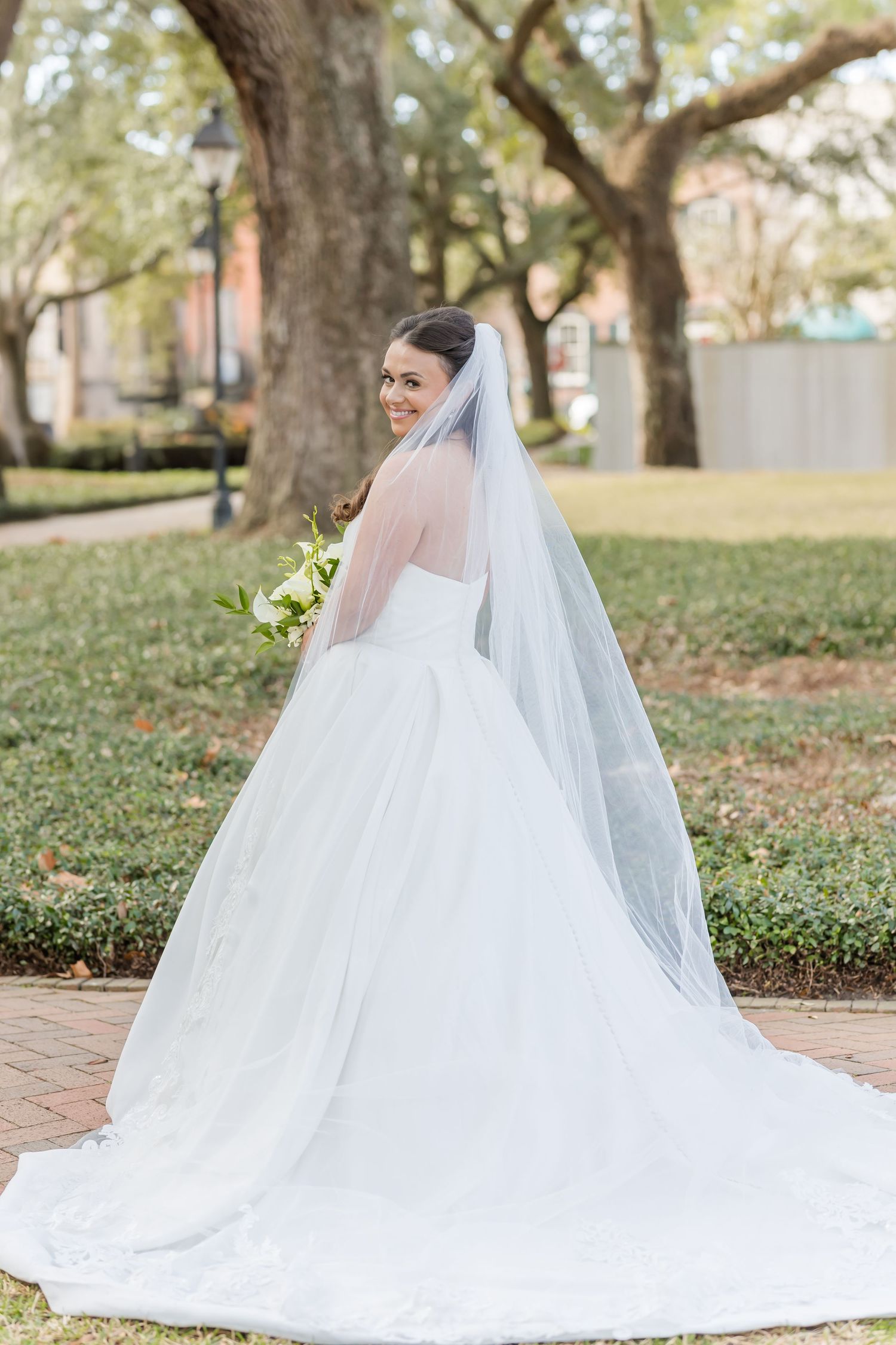 Bride in a flowing white ball gown with long veil turns to smile over her shoulder in a lush outdoor garden setting.