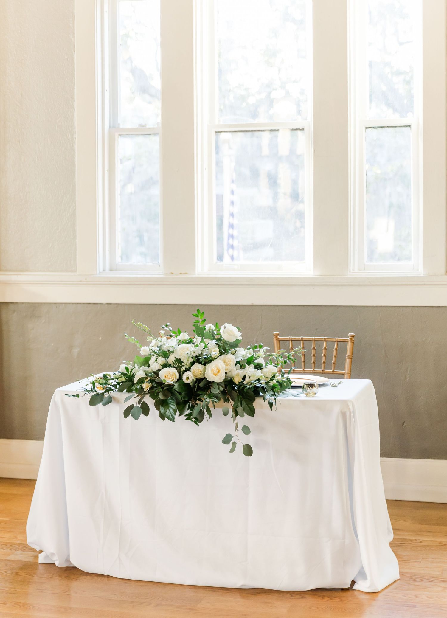Elegant wedding sweetheart table with white linen and lush greenery floral arrangement of white roses and eucalyptus.