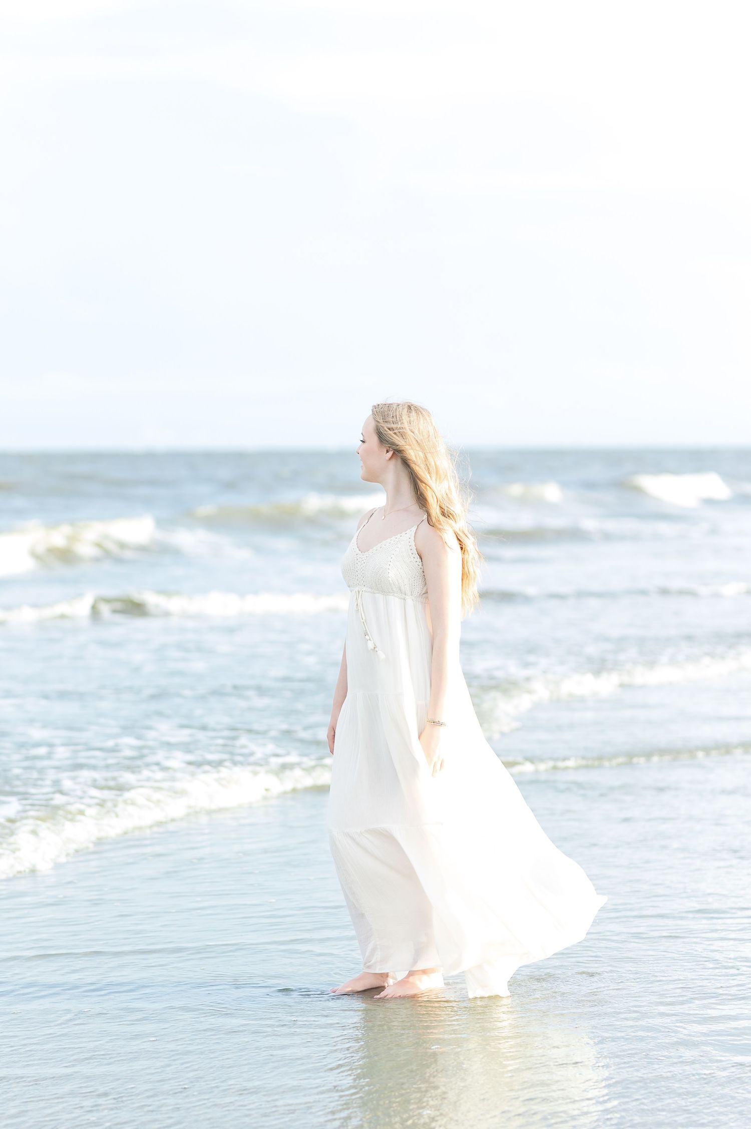 A flowing white dress catches the ocean breeze as a figure stands at the water's edge.