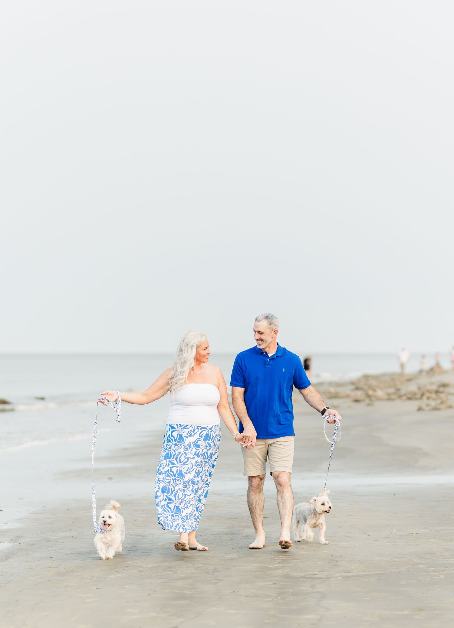 Happy couple walks their two small white dogs along a peaceful beach shoreline on an overcast day.
