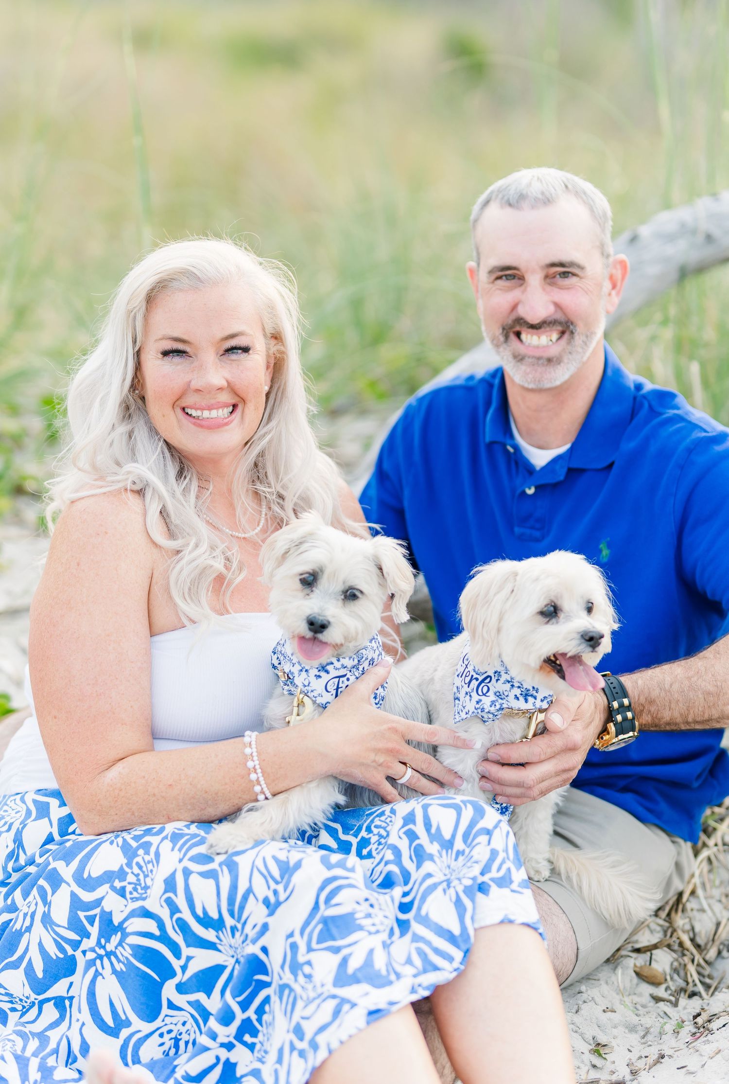 A beachside family portrait with two small white dogs wearing matching blue floral bandanas.
