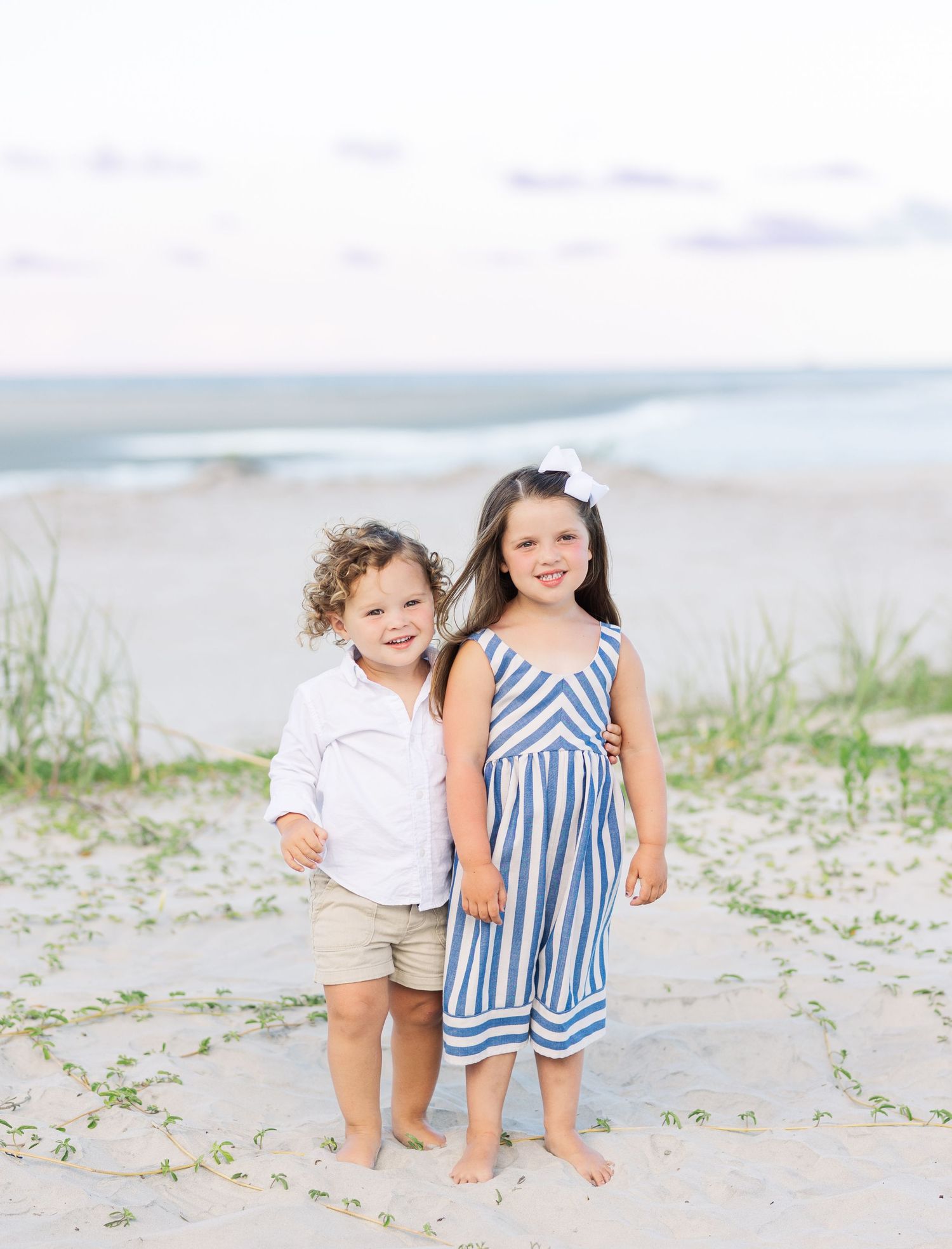 Two children stand together on a sandy beach with ocean waves in the background on a bright sunny day.