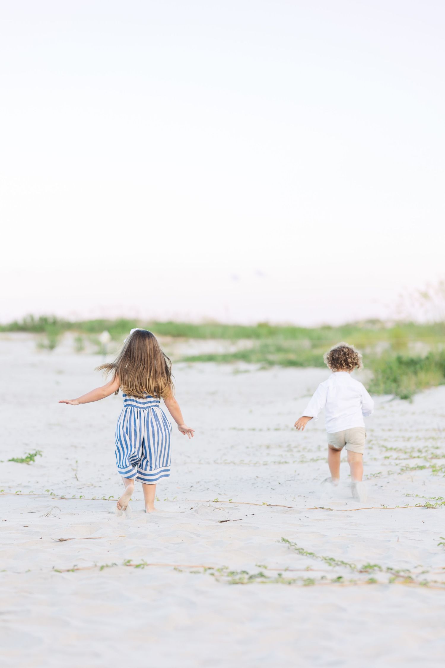 Two children run playfully along a sandy beach with one wearing a striped dress and the other in white.