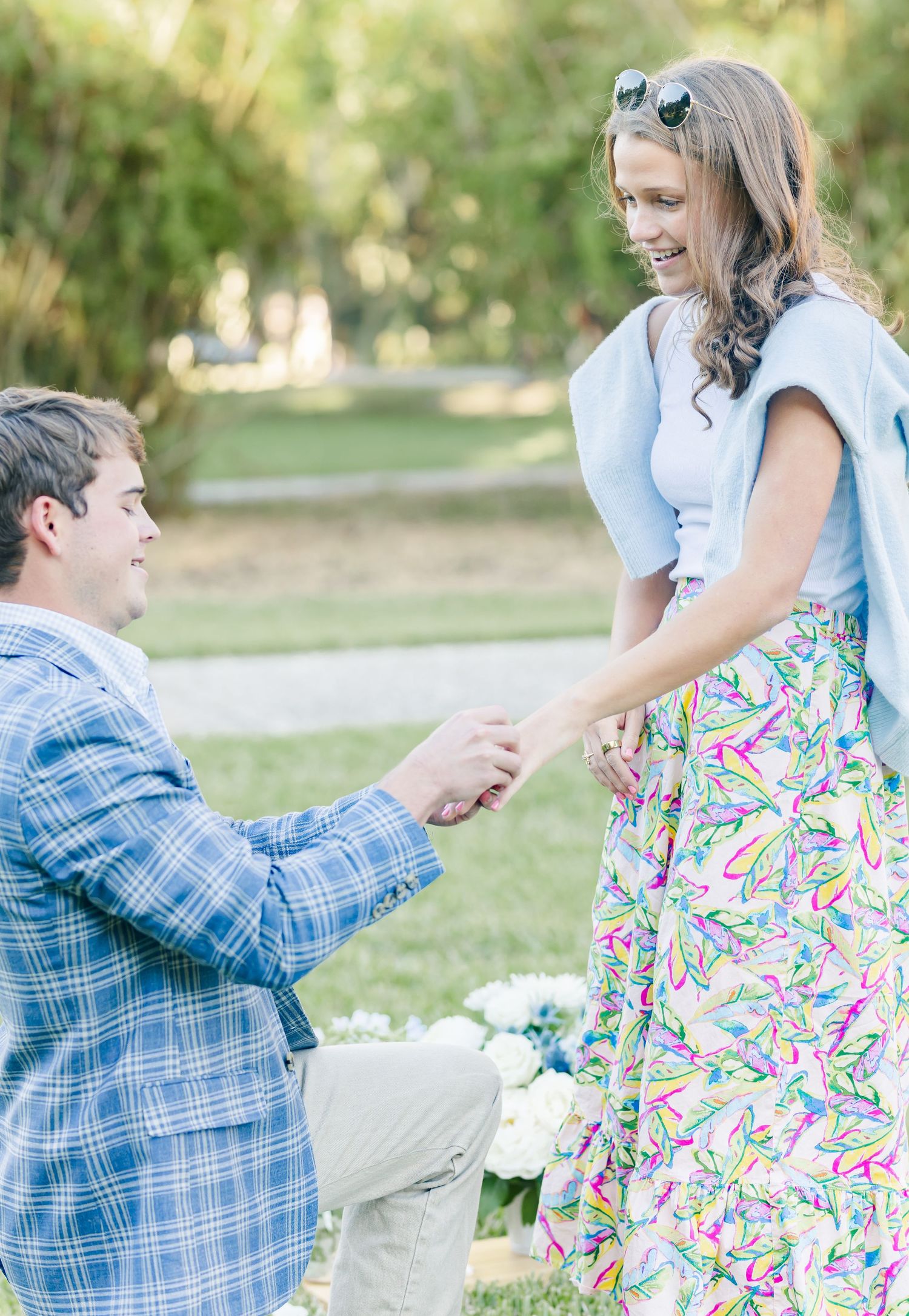 A romantic proposal scene unfolds in a park setting with white flowers on the ground.
