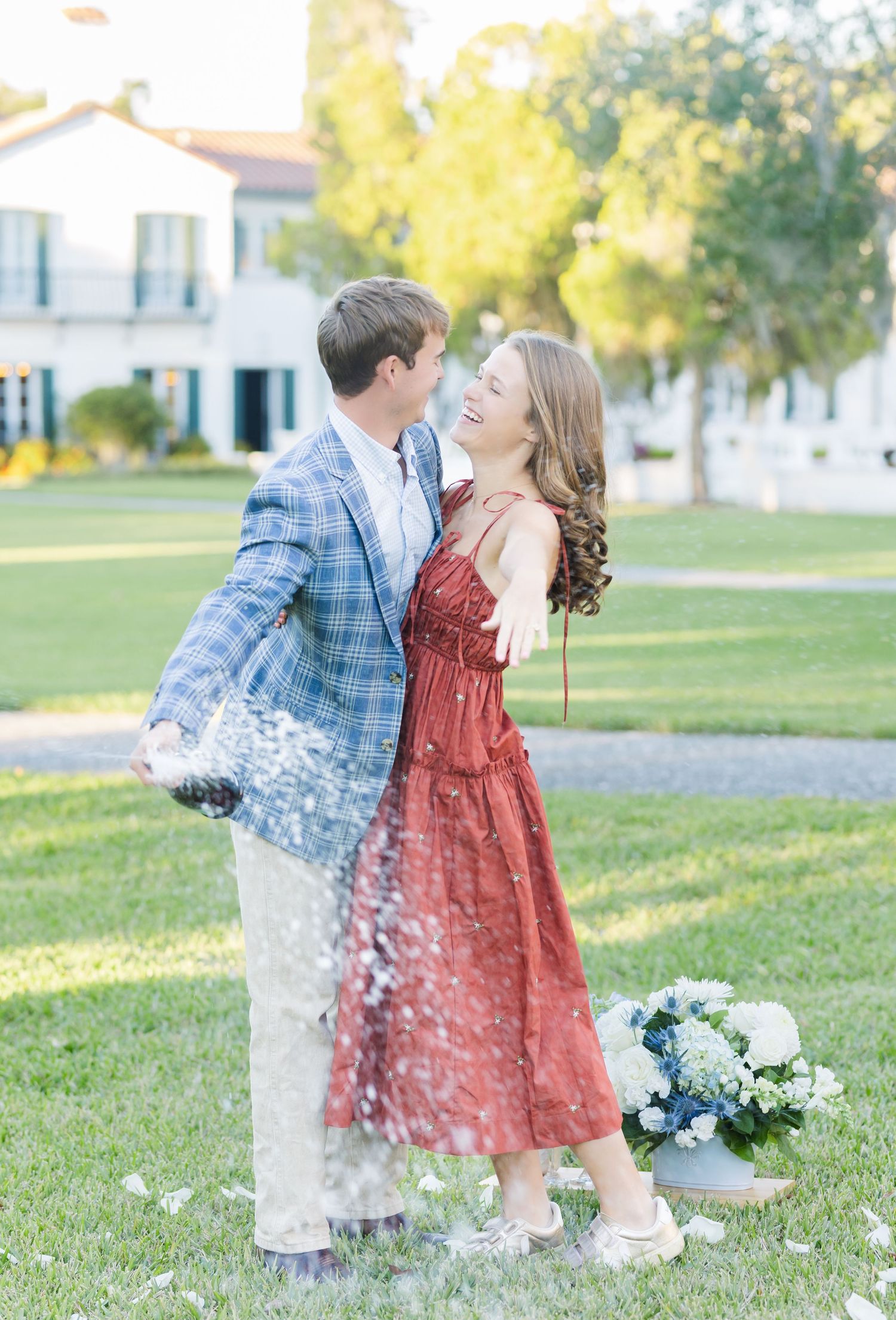 A couple in blue plaid and red dress playfully spray champagne in a sunny outdoor setting.
