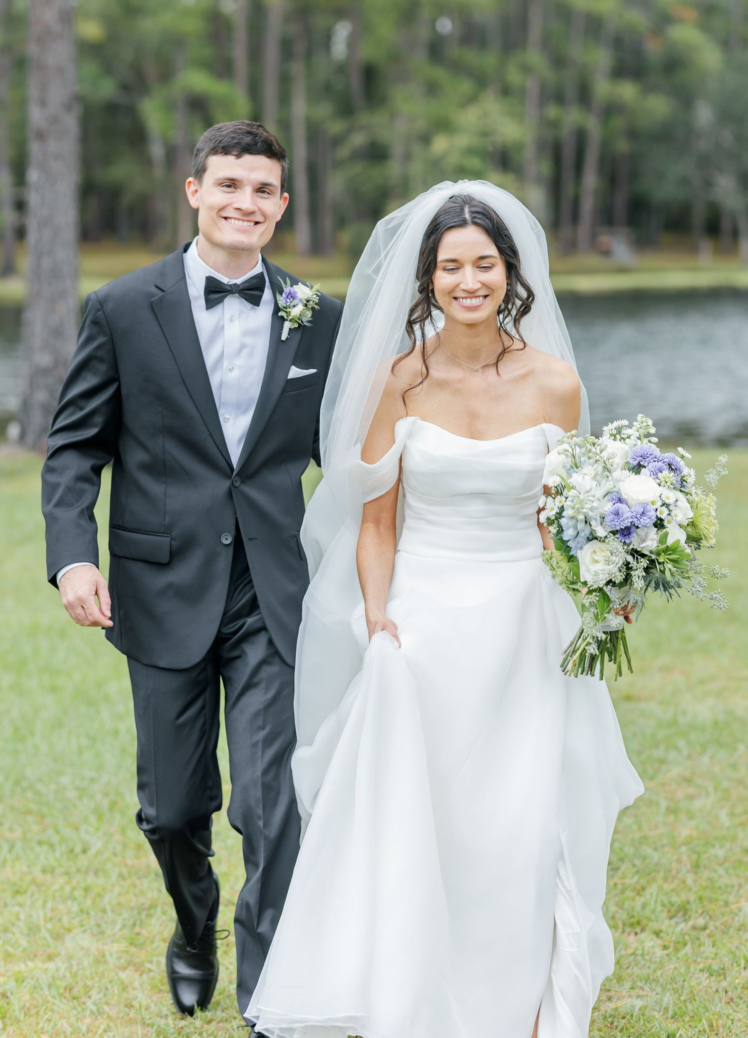 A wedding couple poses together outdoors by a lake with a beautiful floral bouquet.