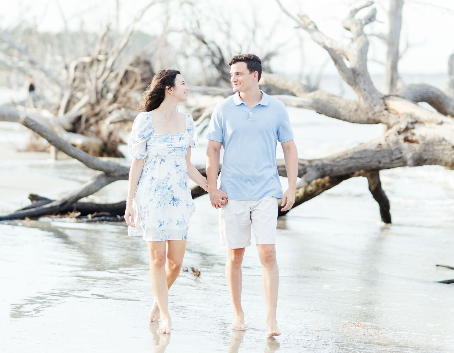 Couple walking hand in hand along a serene beach with driftwood, wearing light blue and floral summer attire.